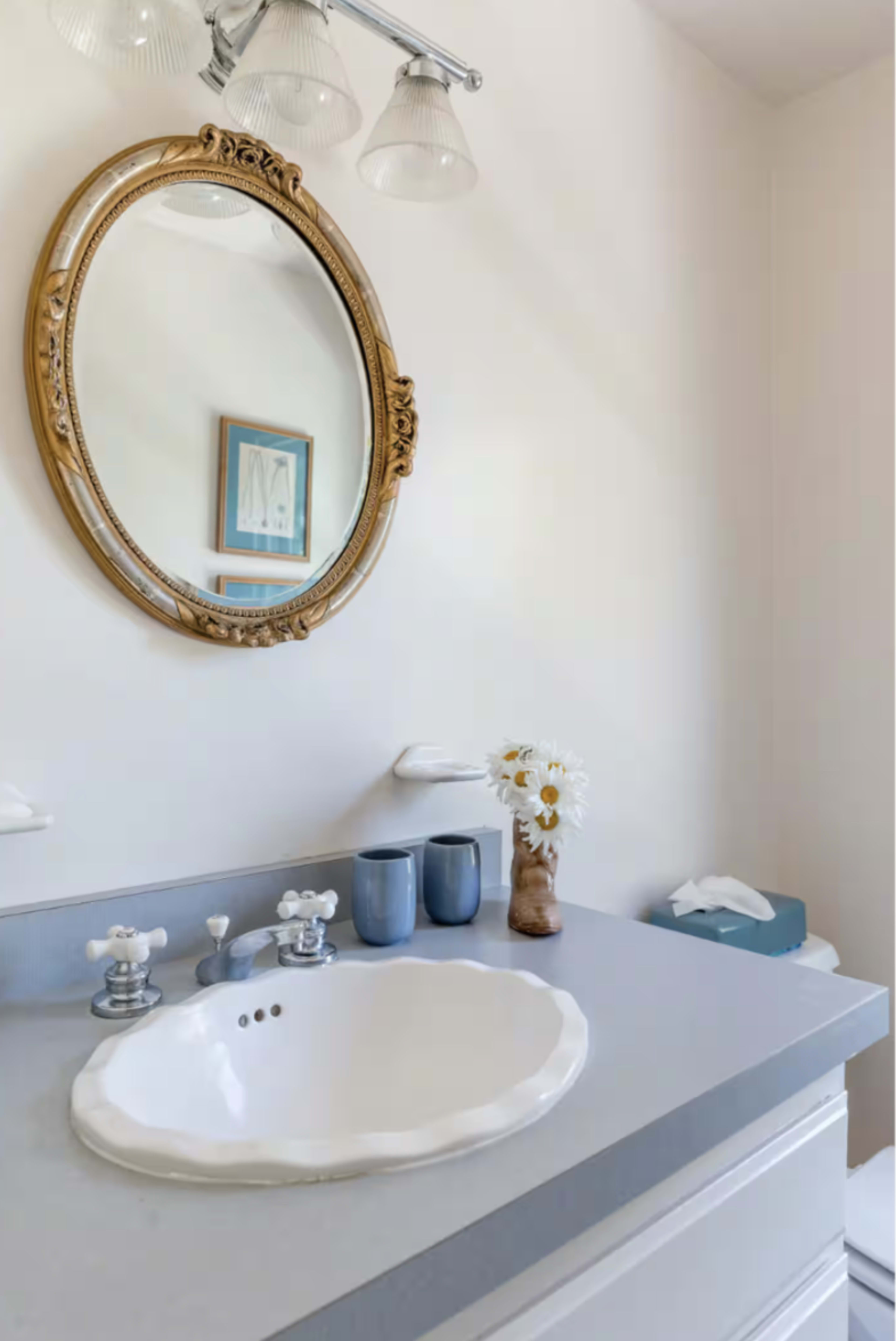 A bathroom features a circular mirror above a sink with a decorative gray countertop, flanked by two blue vases and a framed artwork on the wall.