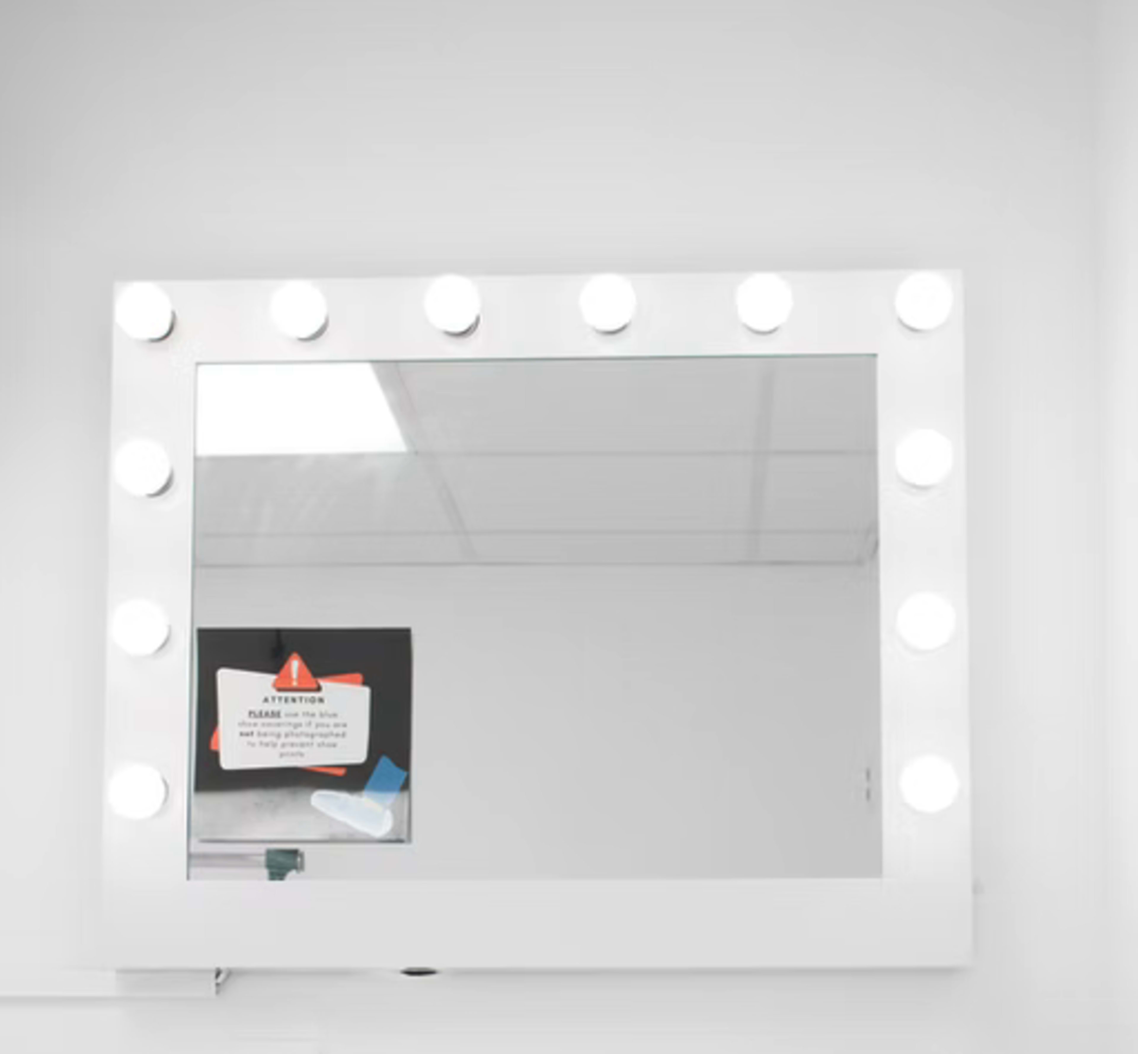 The image shows a well-lit makeup mirror with bright bulbs surrounding it, mounted on a white wall, reflecting a printed notice in the background.