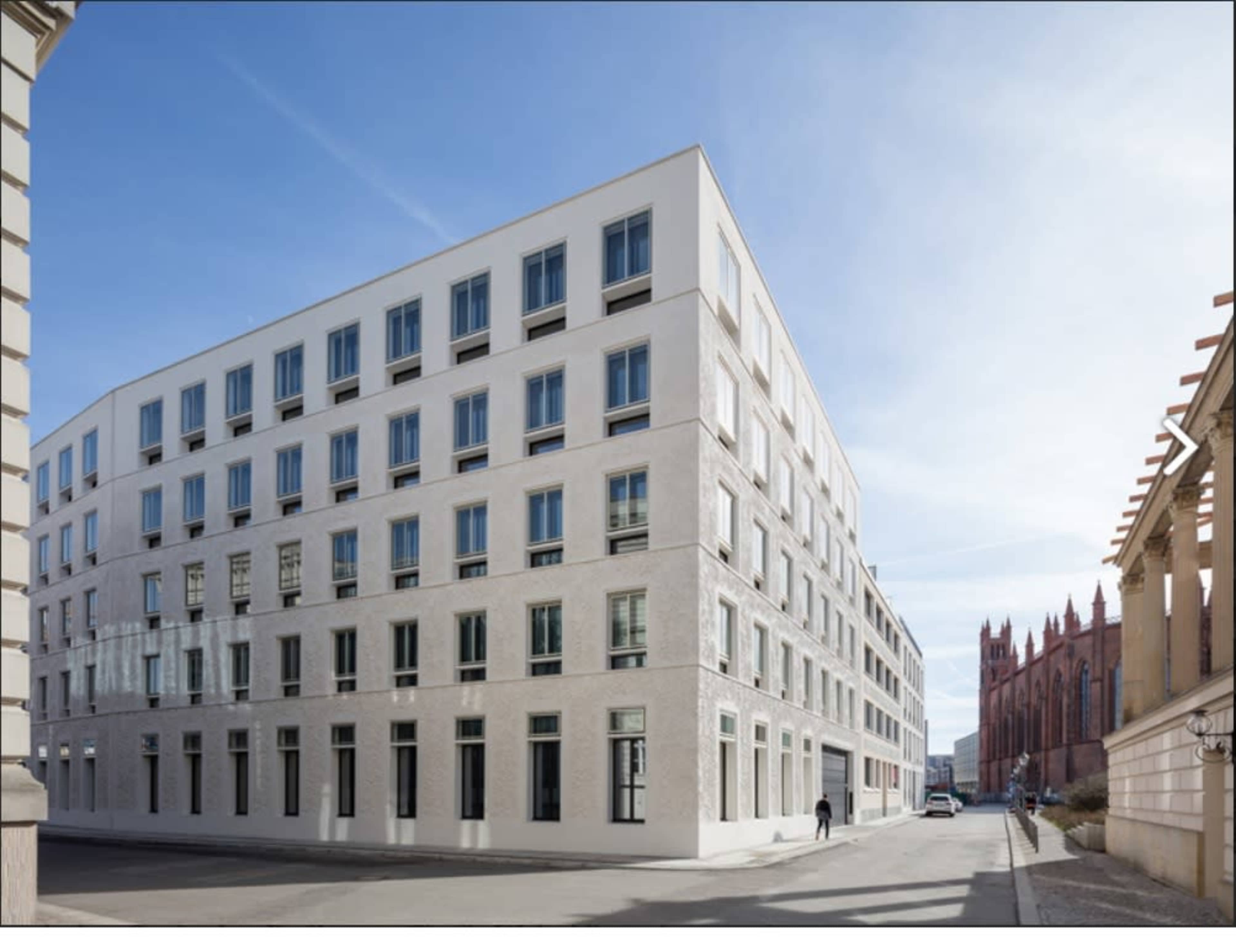 A modern, four-story building with a white facade stands at the corner of a street, adjacent to a historic red-brick structure.