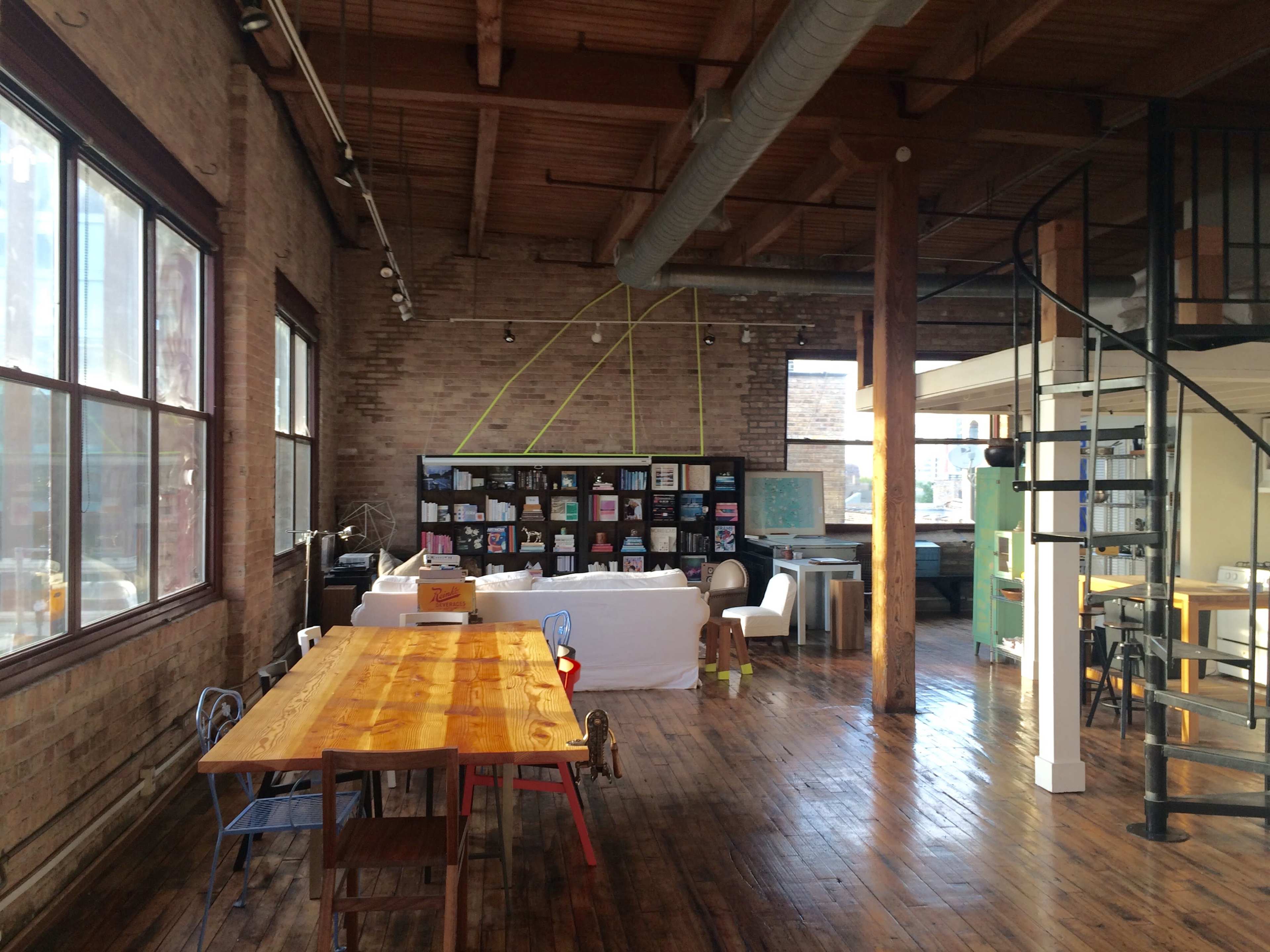 A spacious, industrial-style room with large windows, wooden flooring, and a long dining table, along with various furniture pieces and a spiral staircase.
