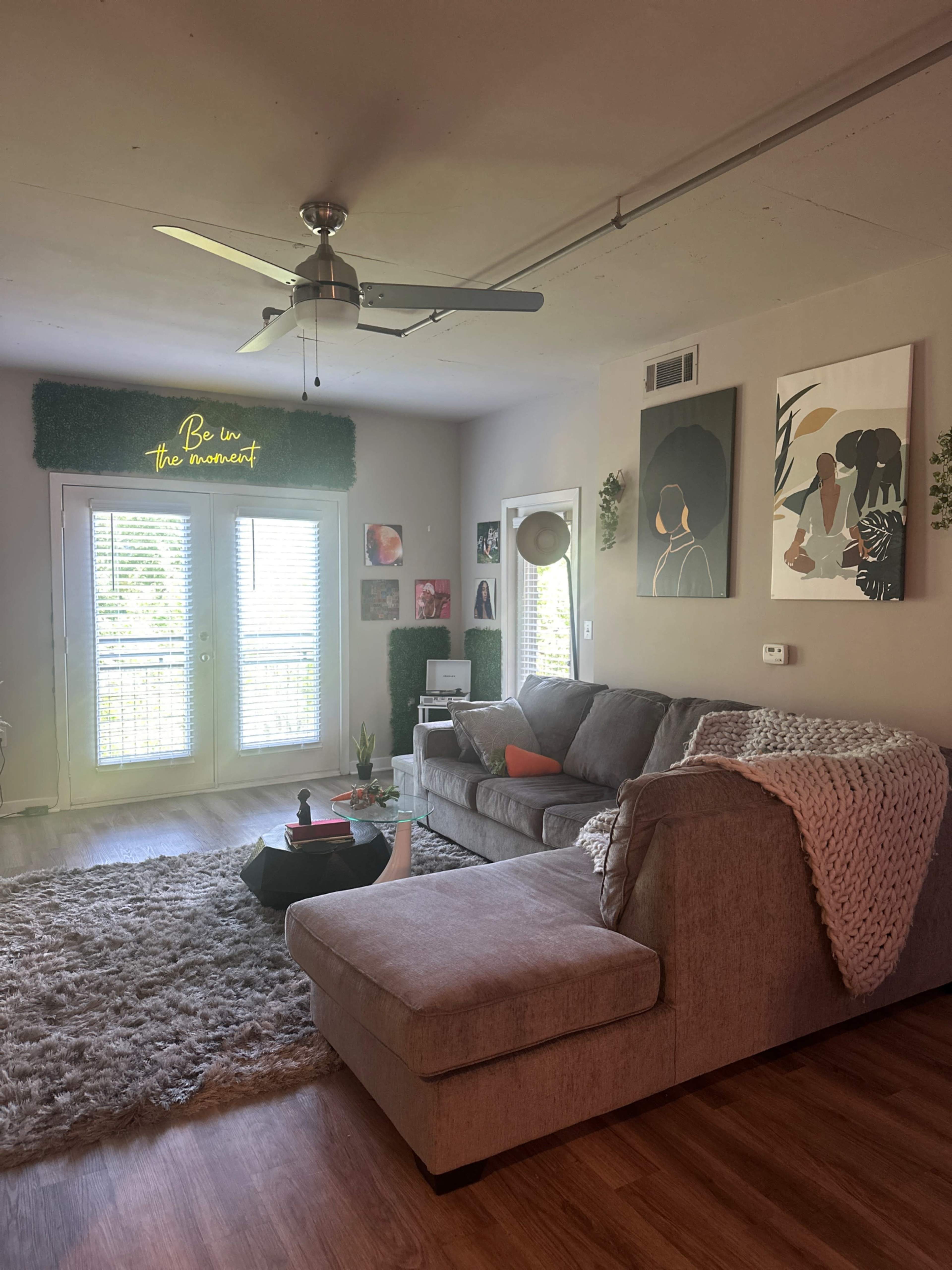 The image shows a cozy living room with a gray sectional sofa, decorative wall art, and a large window leading to a balcony.