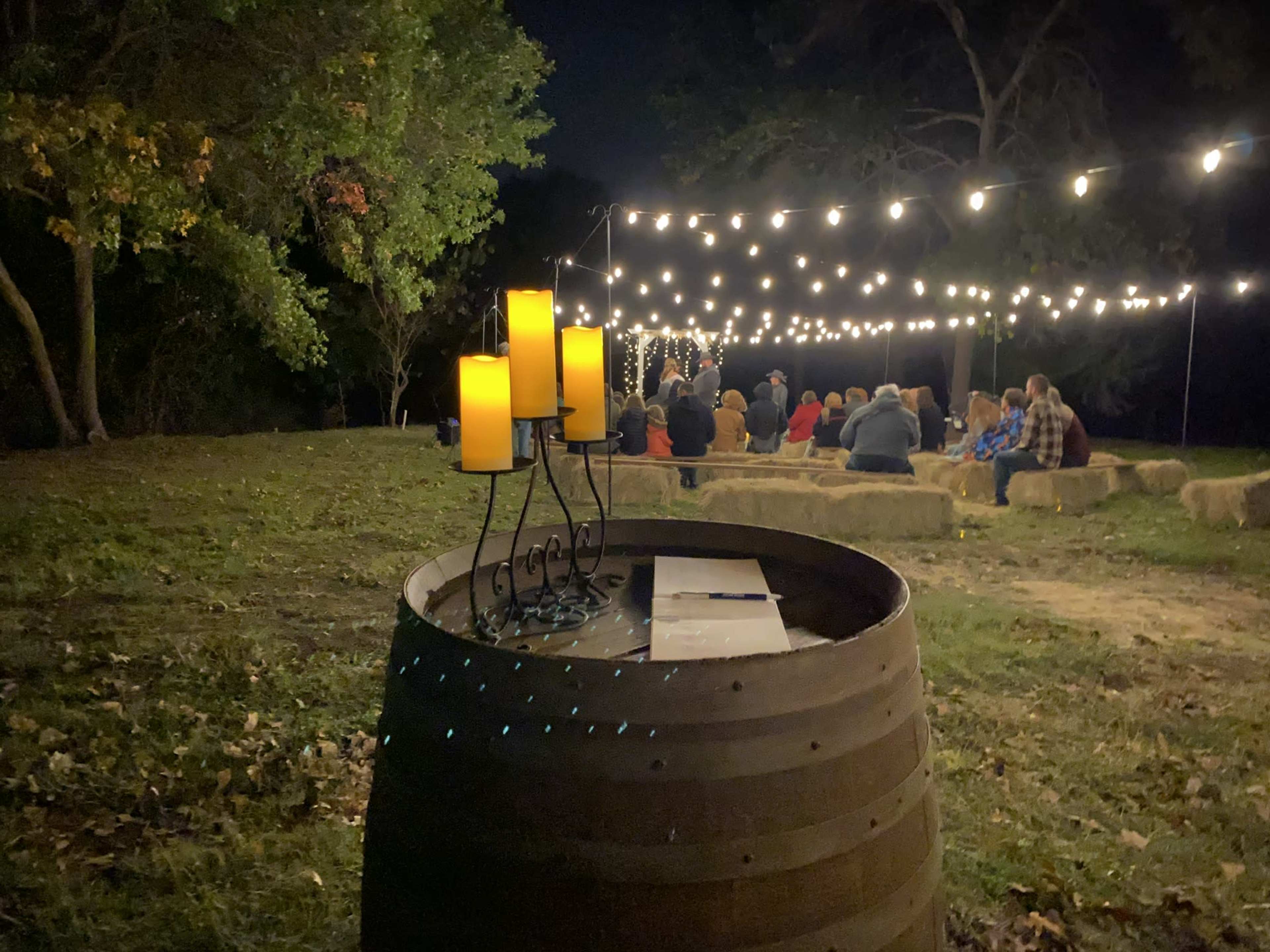 A group of people sits in a grassy area beneath strings of lights, while candles are arranged on a barrel in the foreground.