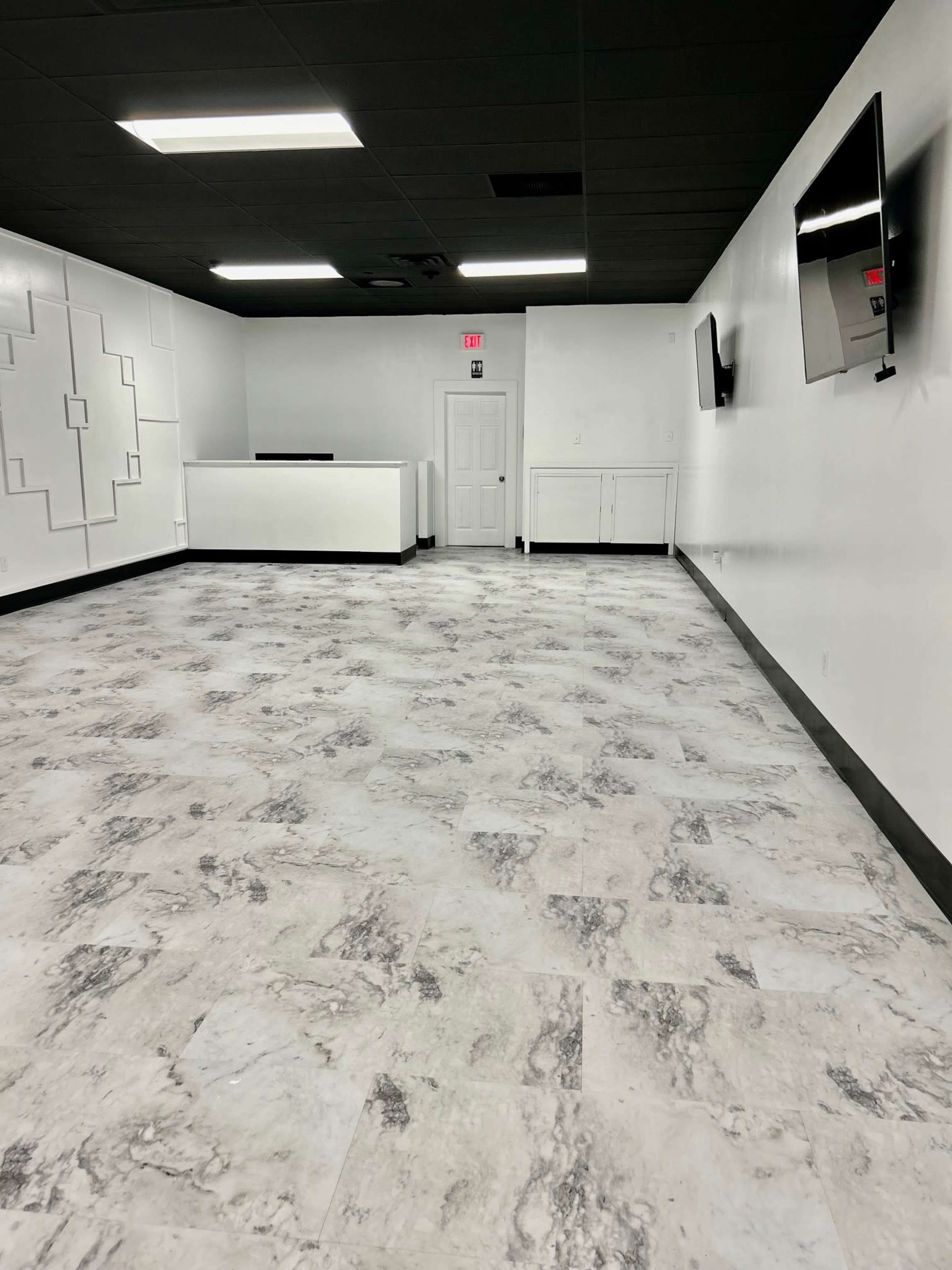 An empty, well-lit room with marble-patterned flooring, a white counter in the back, and two wall-mounted televisions.