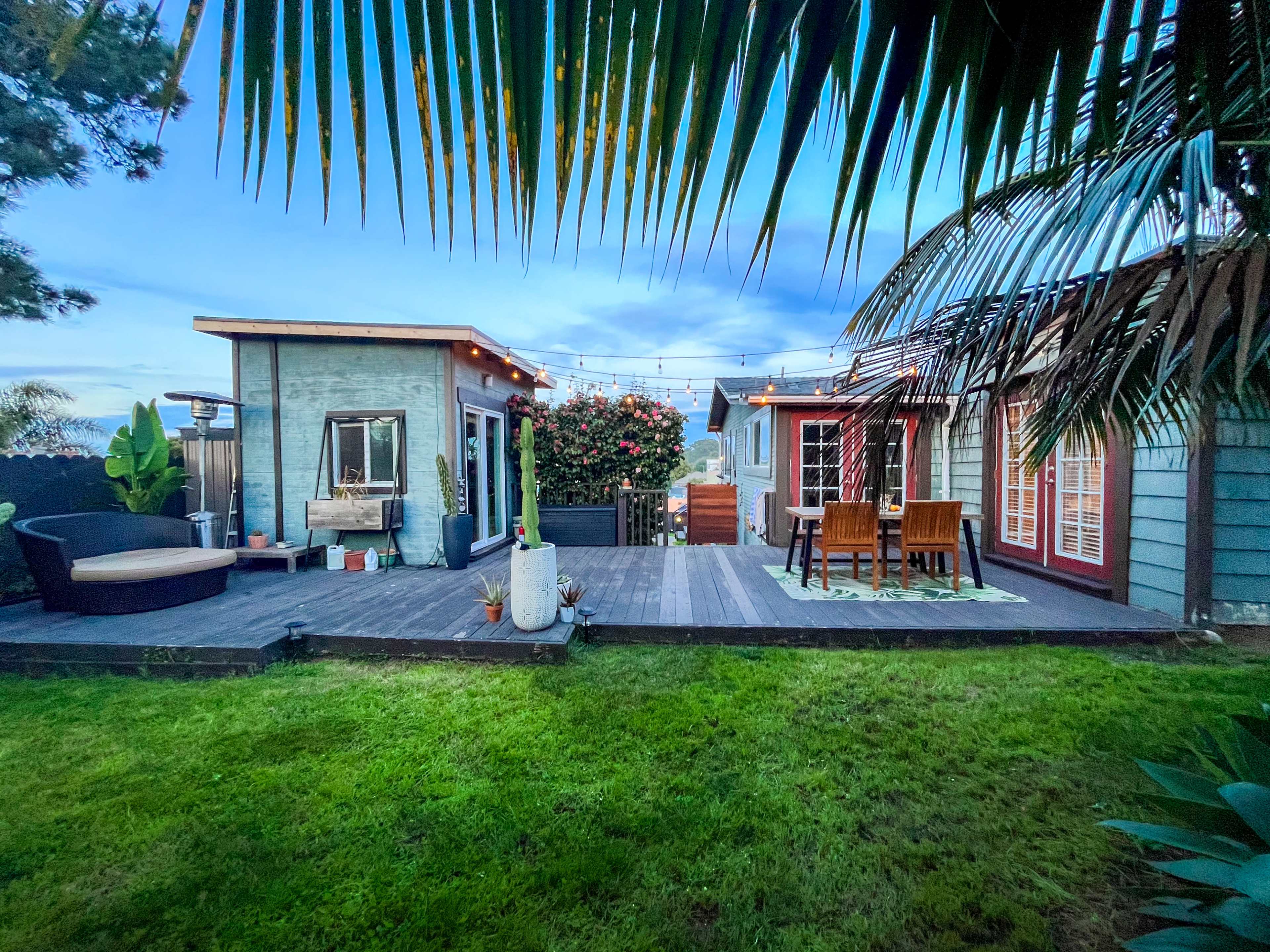 A backyard scene features a deck with seating areas, surrounded by greenery and string lights draped above the space.