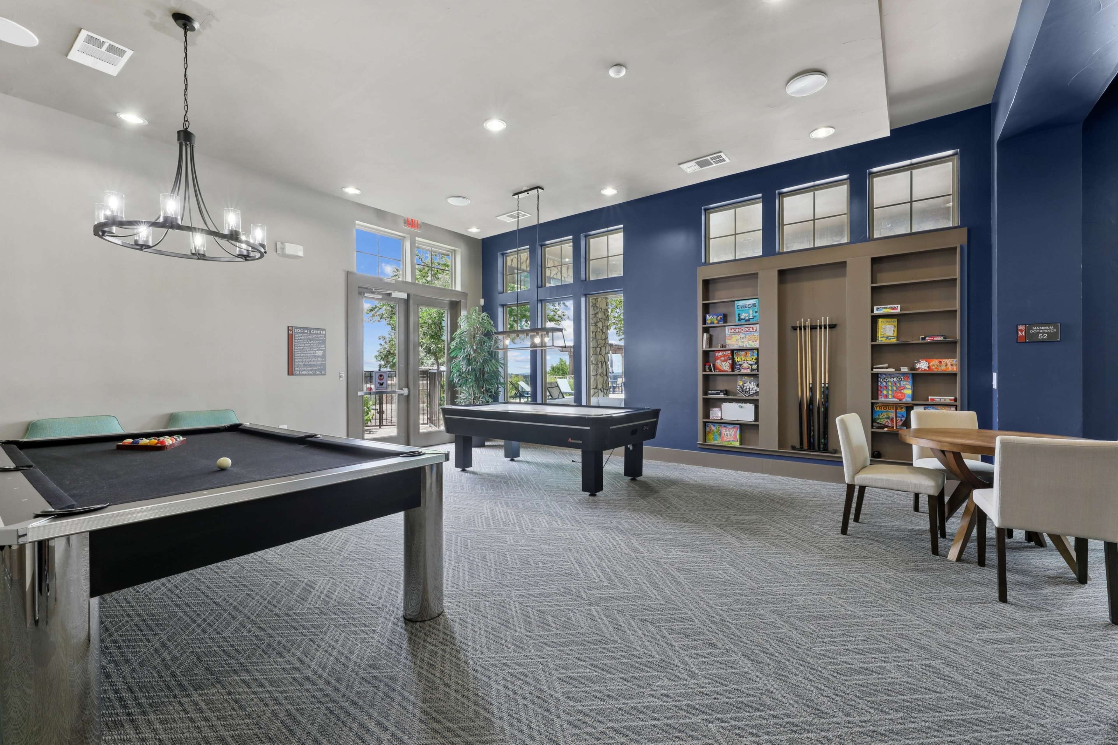 The image shows a recreational room with two pool tables, a dining area, and shelves filled with games, all set against a bright blue wall.