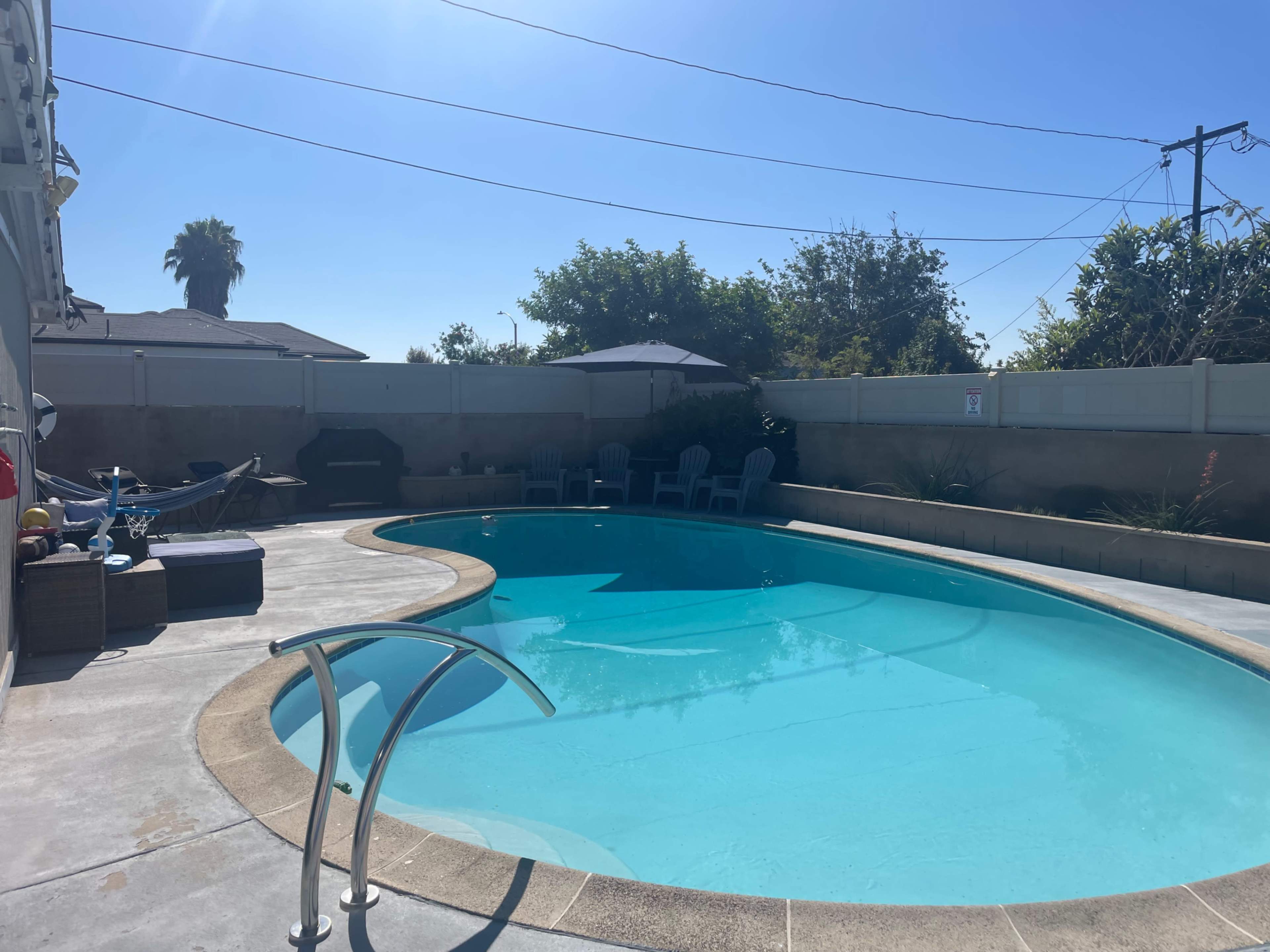The image shows a clear blue swimming pool surrounded by a concrete deck and several chairs, with a palm tree in the background.