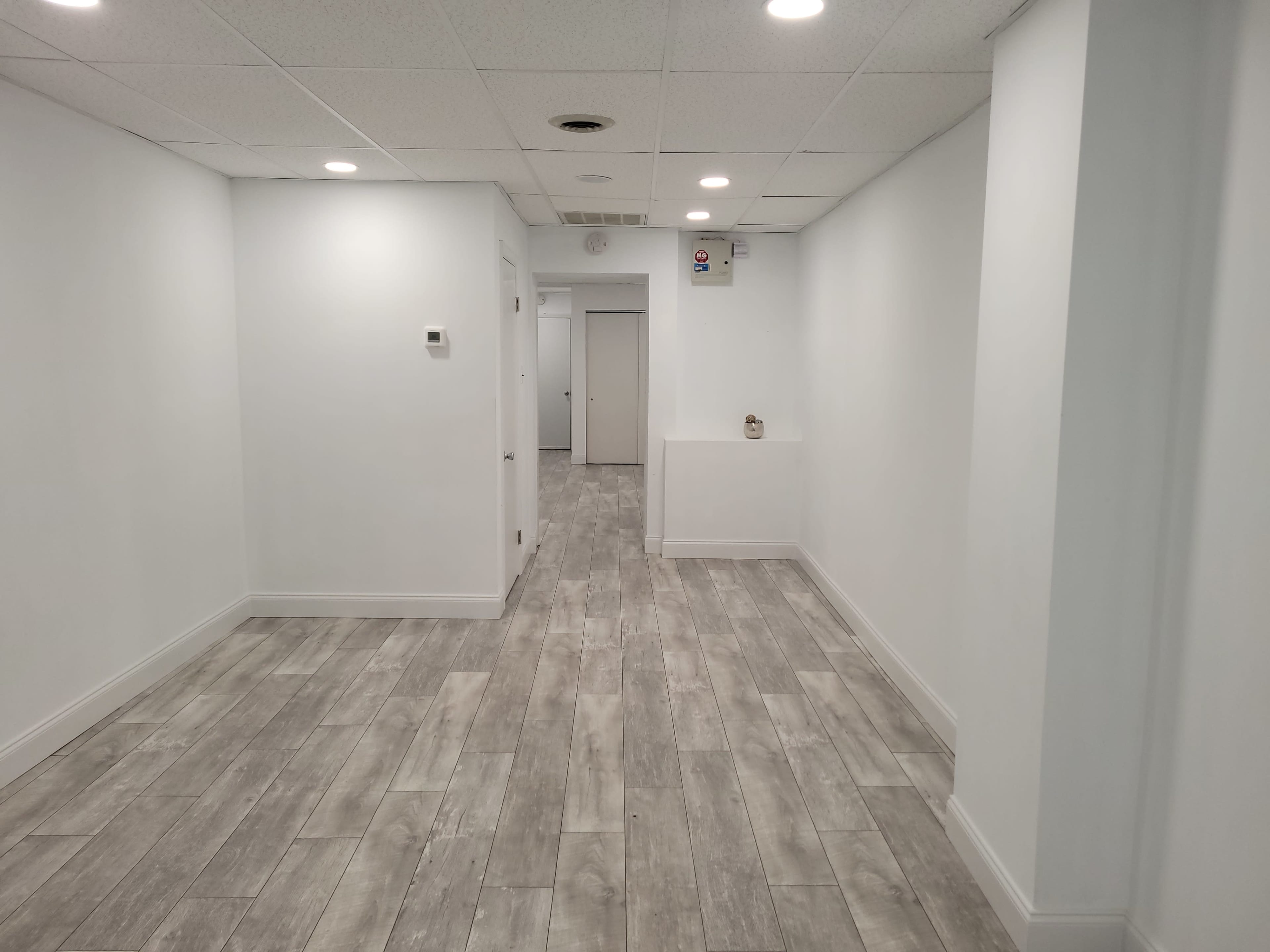 The image shows a long, narrow hallway with white walls, tile flooring, and recessed lighting.
