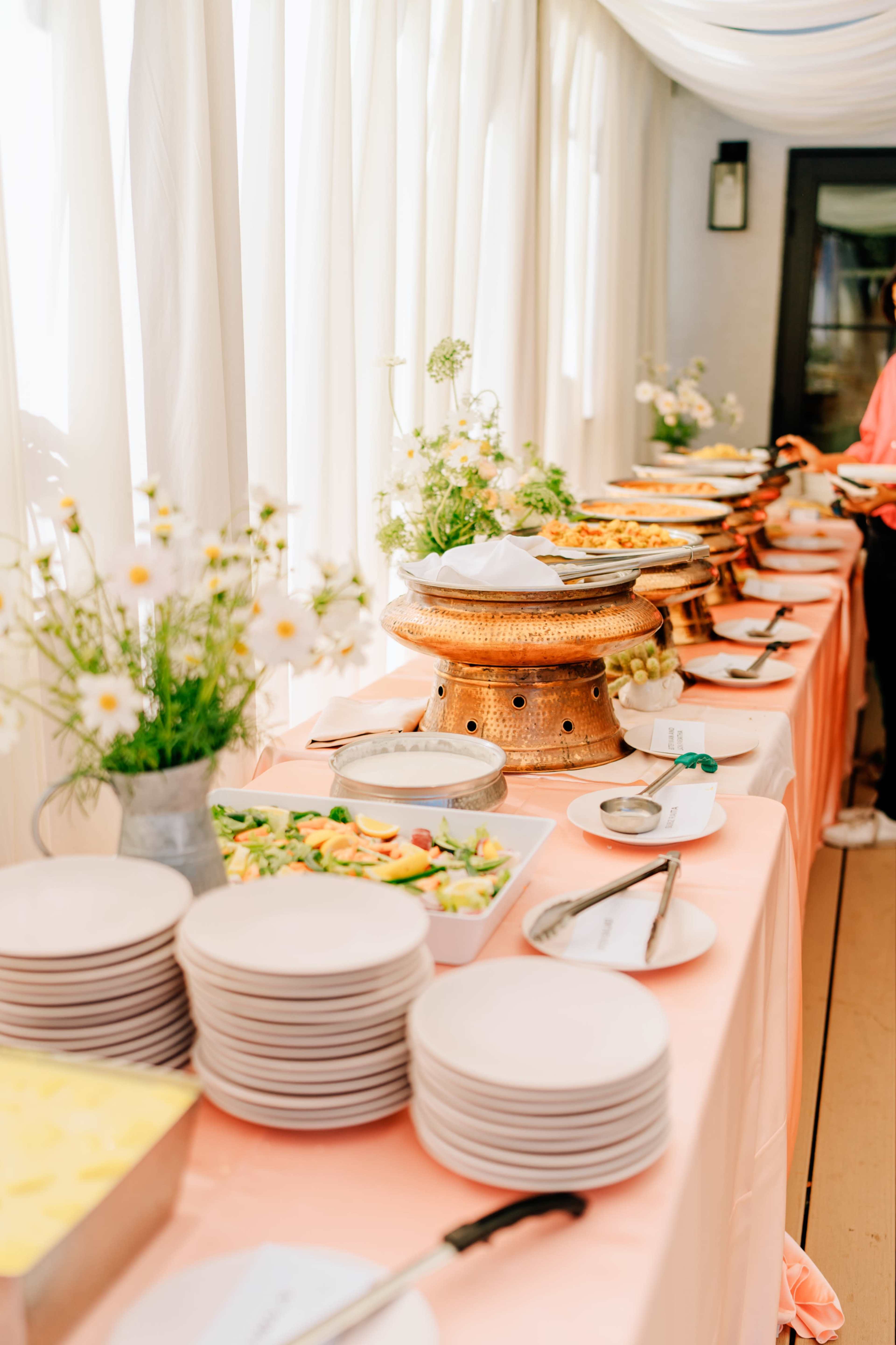 A long table draped in a peach tablecloth displays various dishes in serving bowls, surrounded by plates and decorative flower arrangements.