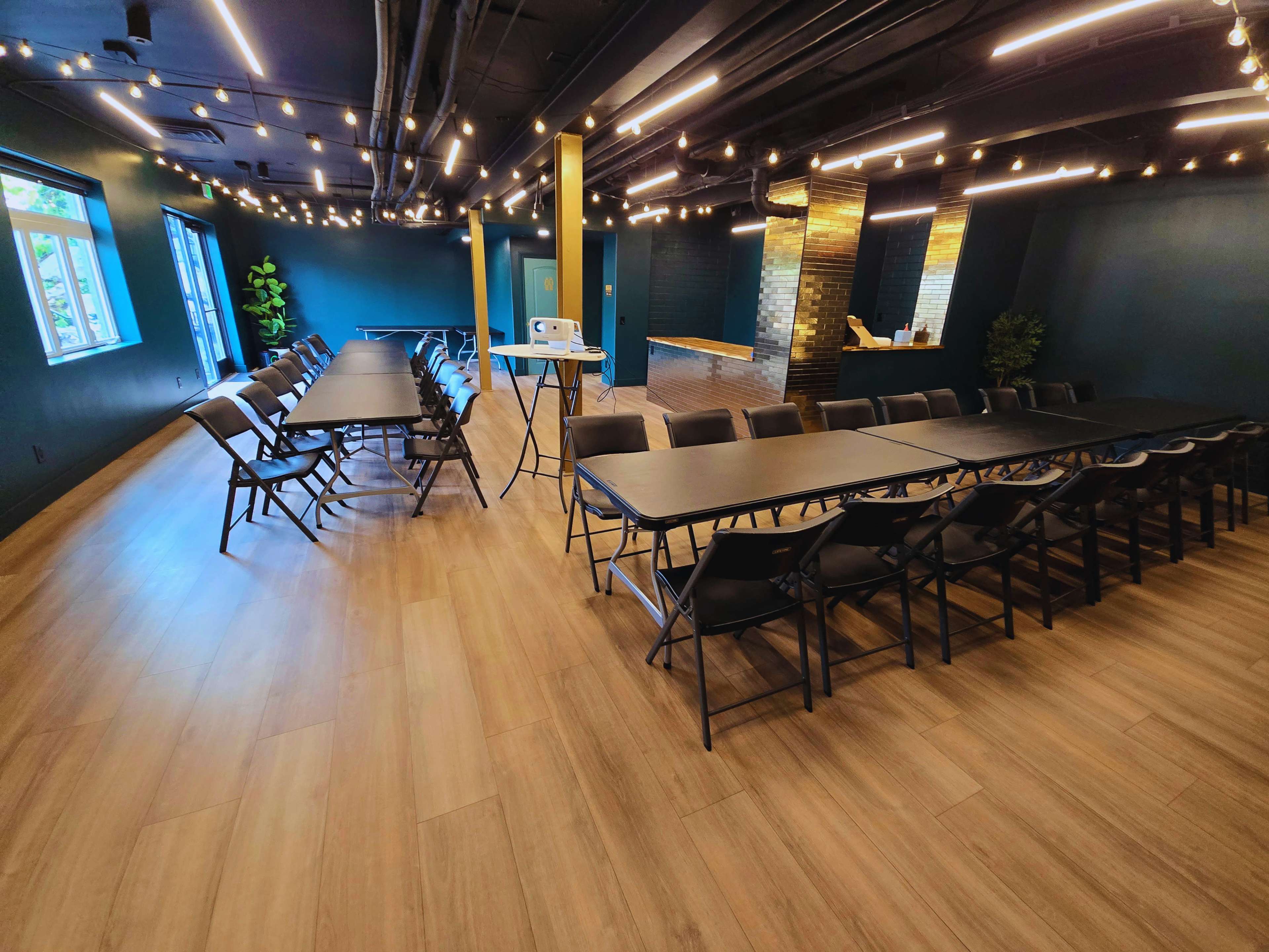 The image shows a large meeting room with multiple tables arranged in a U-shape, surrounded by green walls and warm wood flooring, and illuminated by string lights.