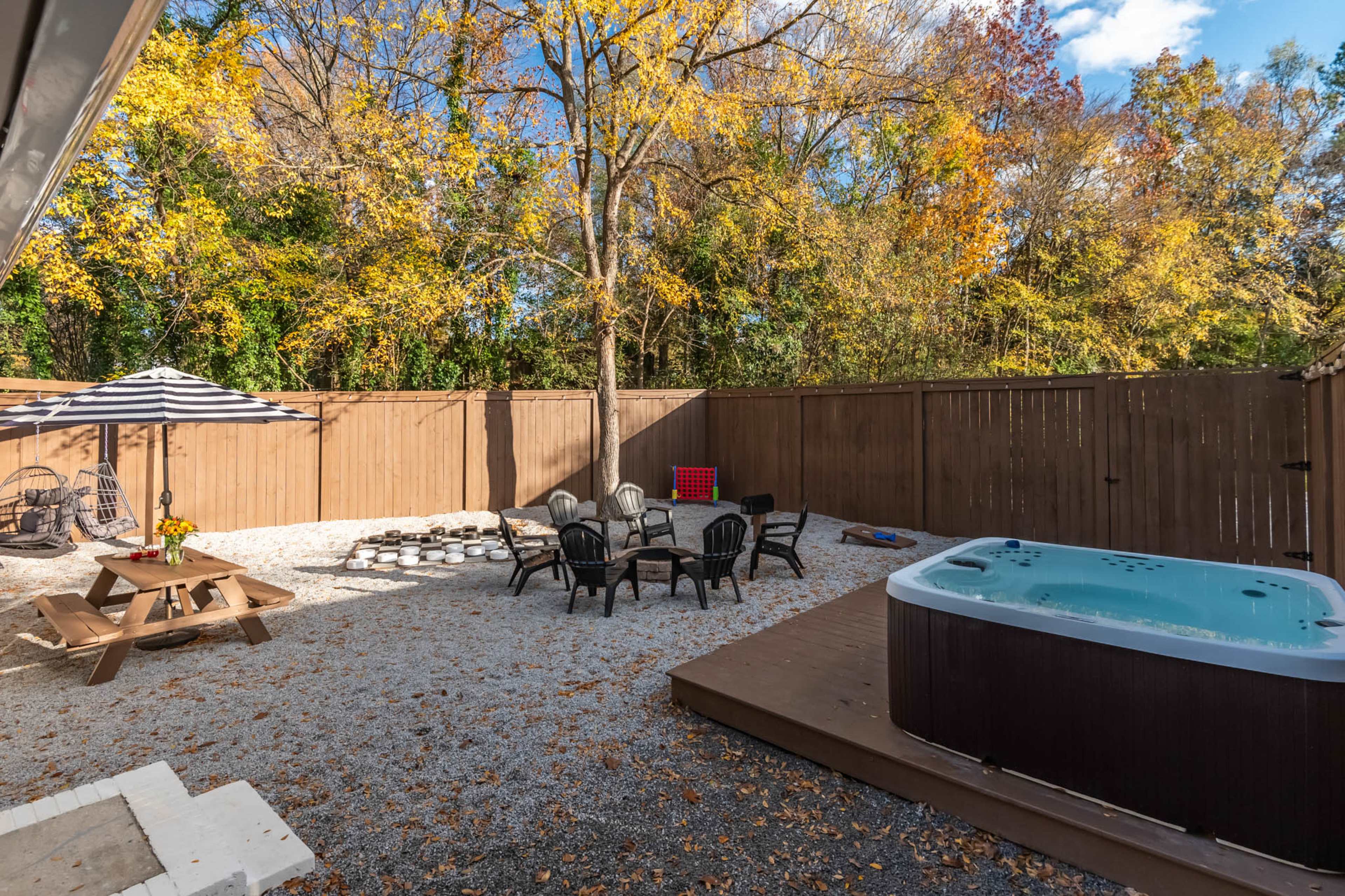 The image shows a backyard with a hot tub, seating area with black chairs, a picnic table, and a shaded patio under a striped umbrella, surrounded by trees in autumn foliage.