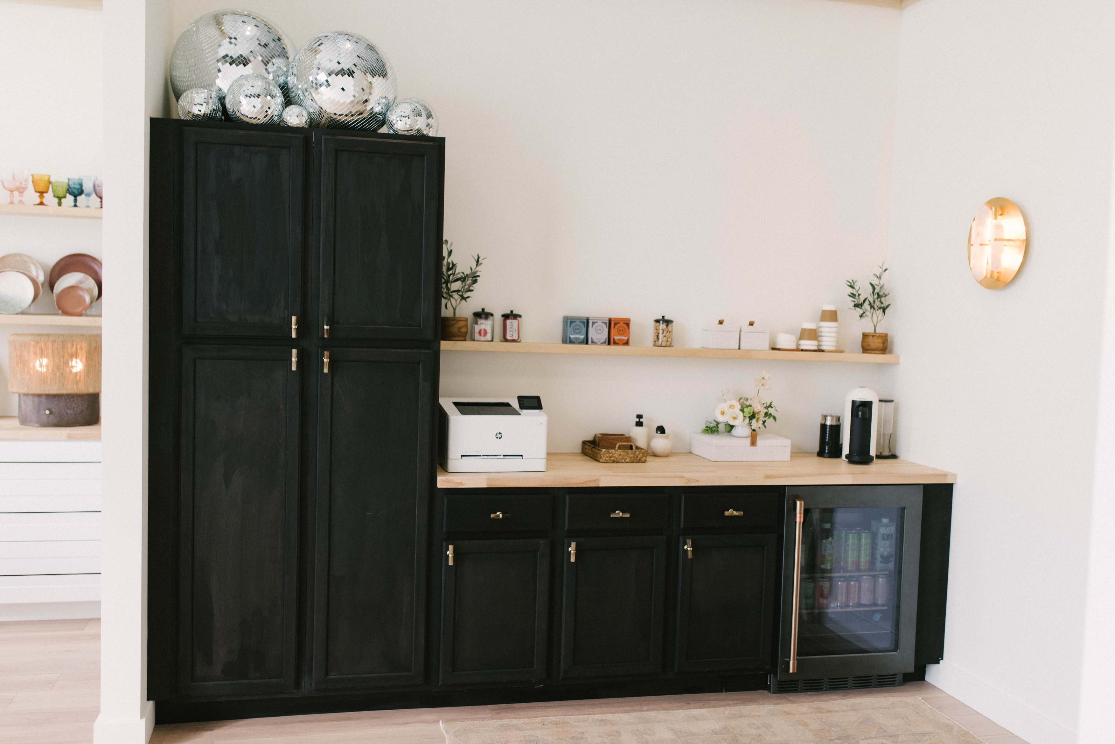 A modern kitchenette featuring black cabinetry, a countertop with decorative items, a printer, and a mini-fridge.