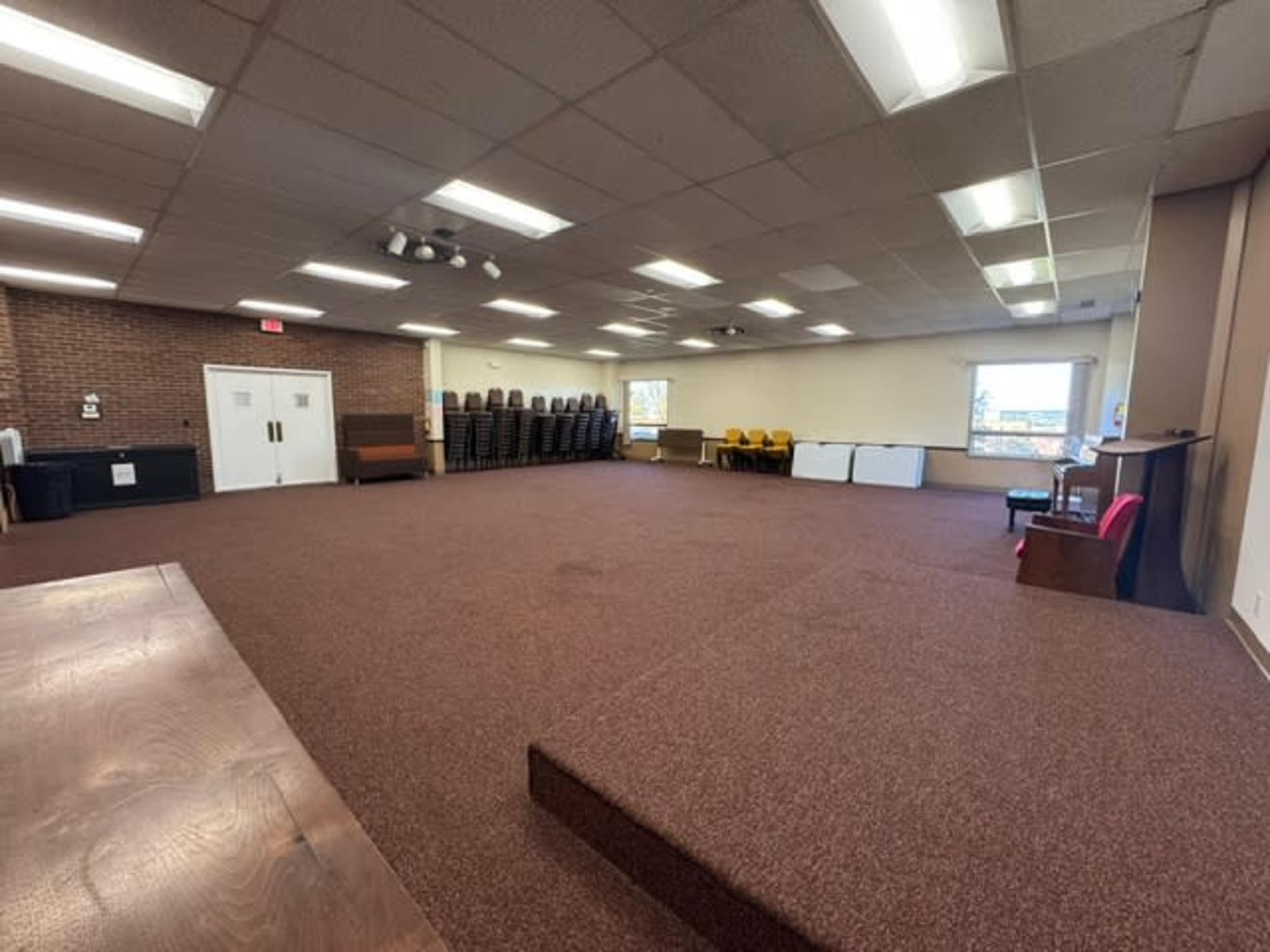 The image shows a spacious room with a carpeted floor, stackable chairs arranged against the wall, and a few pieces of furniture placed around the area.