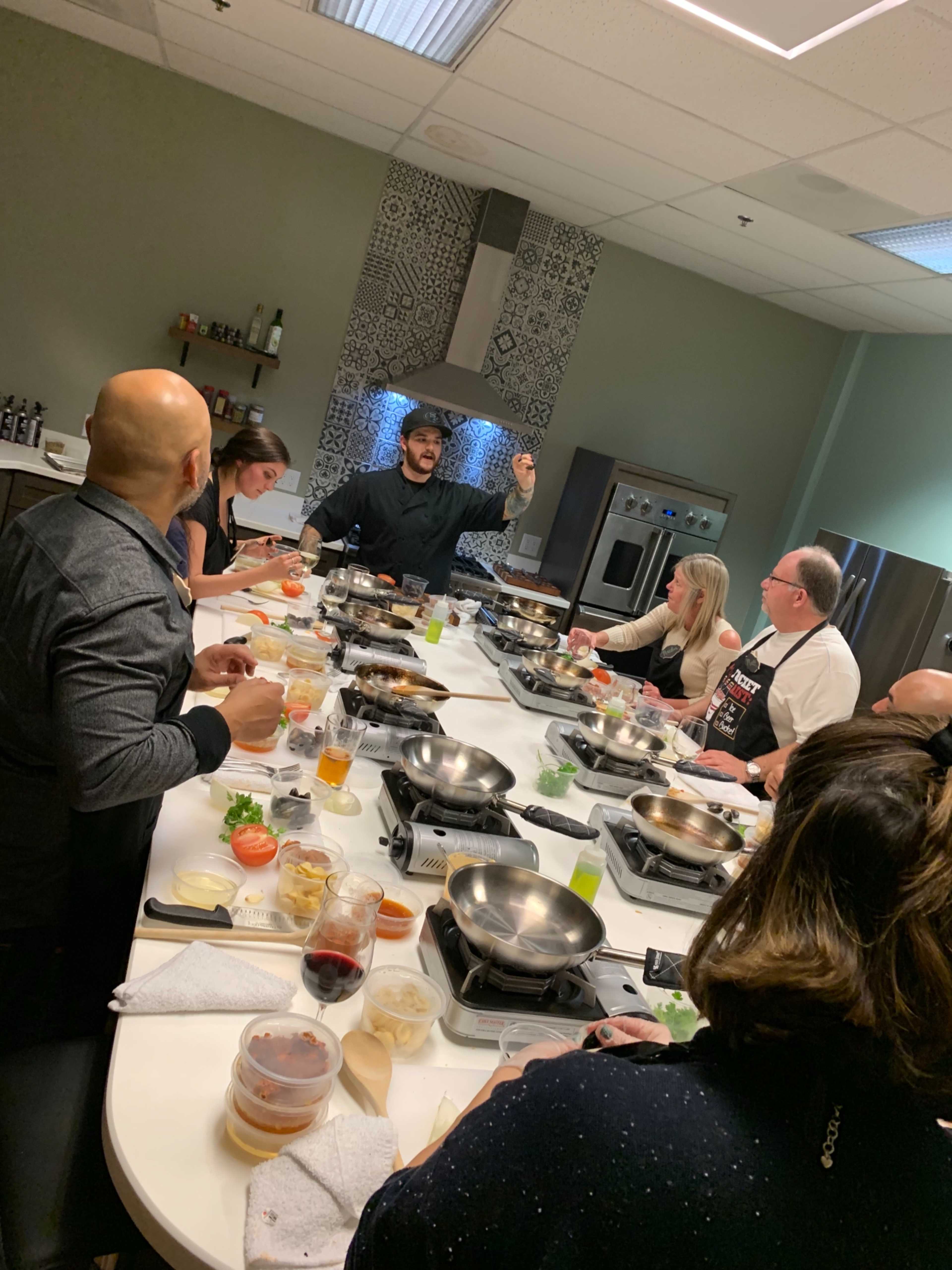 A diverse group of people gathers around a large, circular cooking station as a chef demonstrates a technique in a modern kitchen setting.