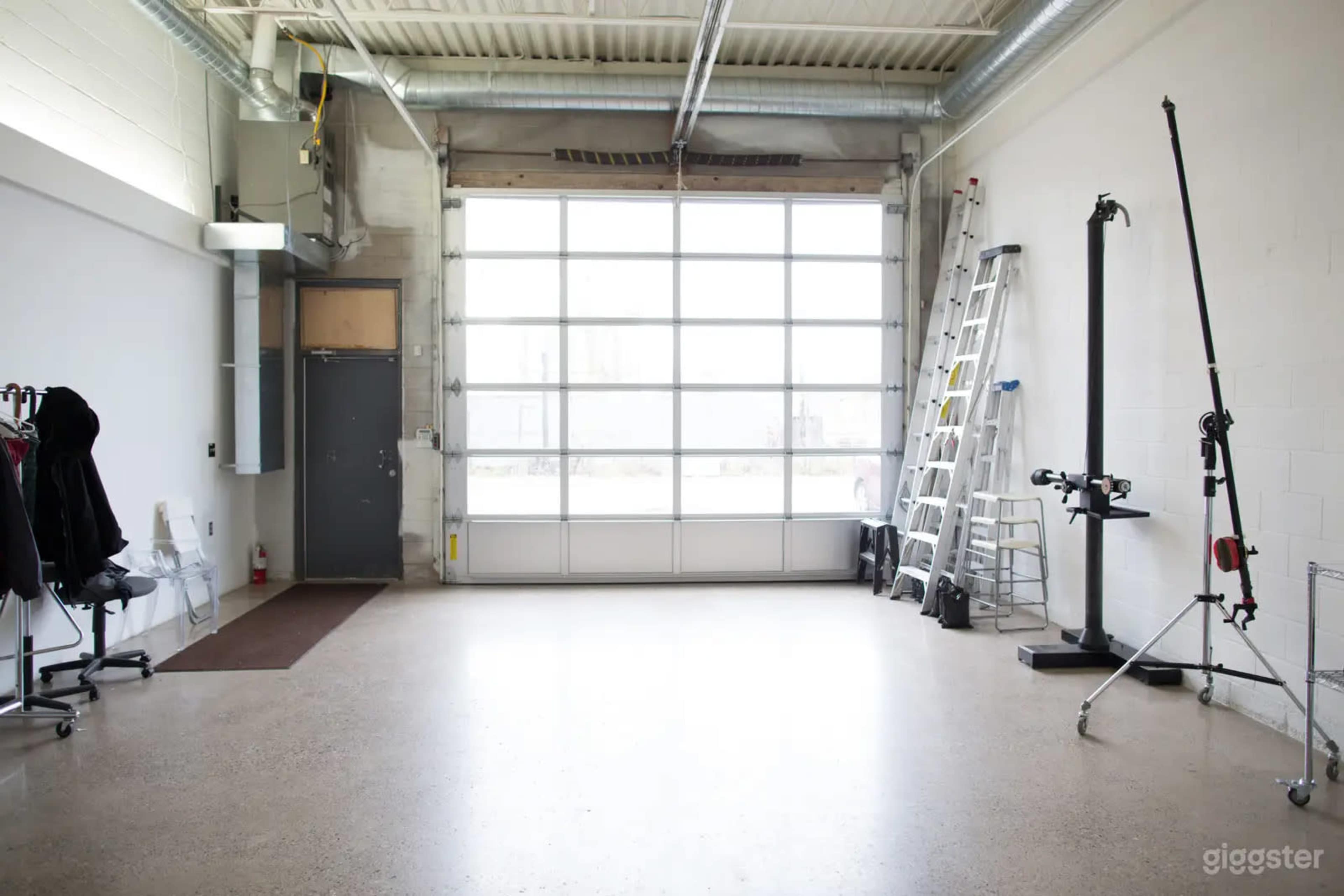 The image shows an empty industrial-style space with a large garage door, a ladder, and some equipment stored along the walls.