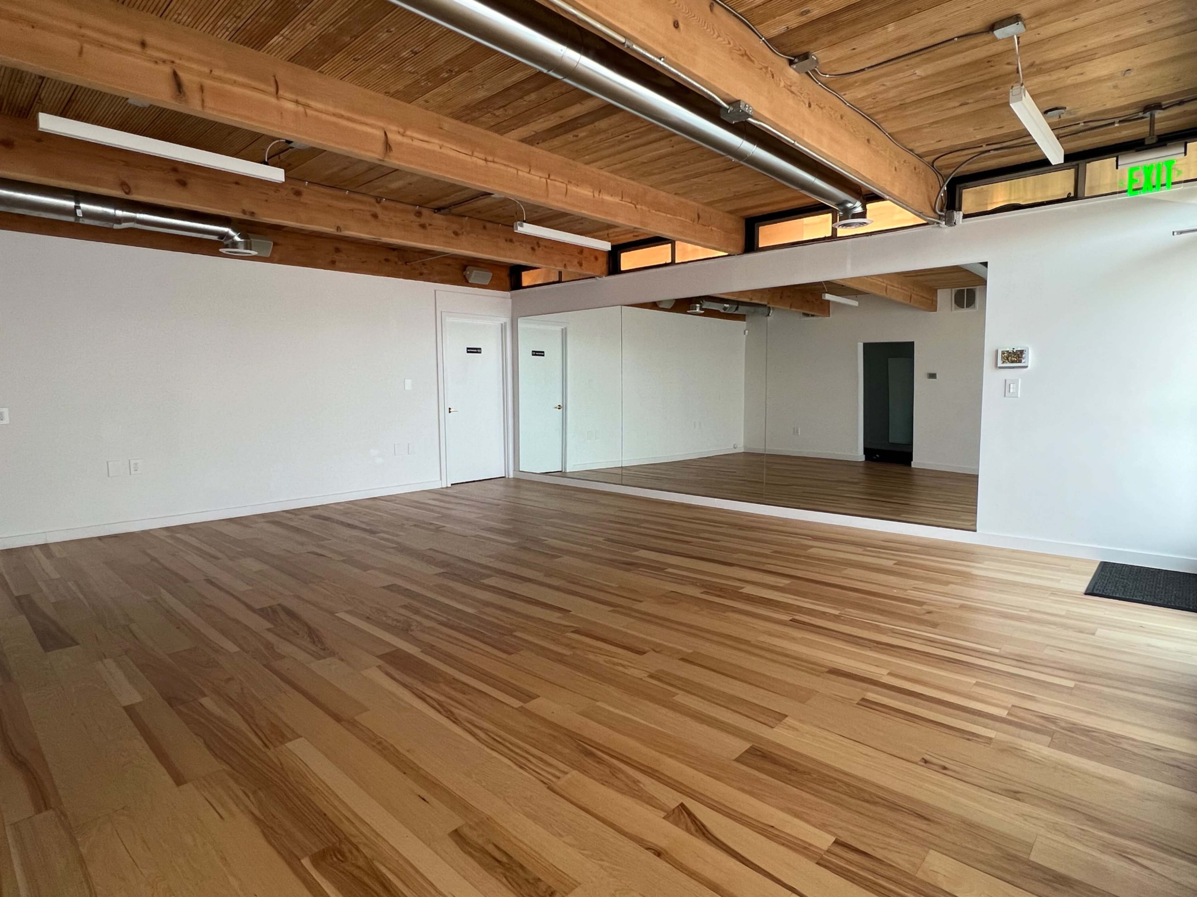 A spacious room with hardwood flooring, exposed wooden beams on the ceiling, and a large mirror on one wall.