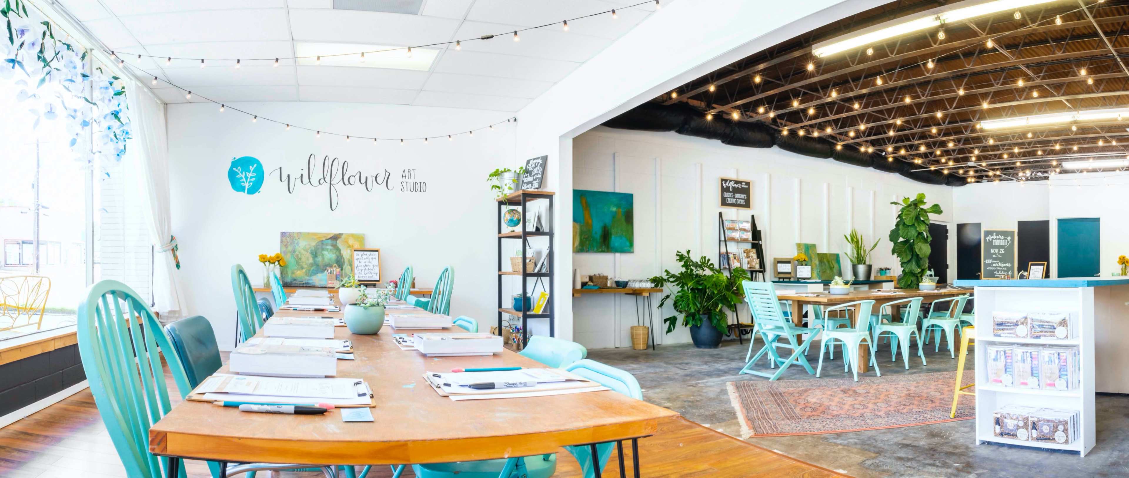 The image shows a spacious art studio featuring a long table set for a workshop, surrounded by plants and colorful chairs, with an inviting atmosphere enhanced by string lights and artwork on the walls.
