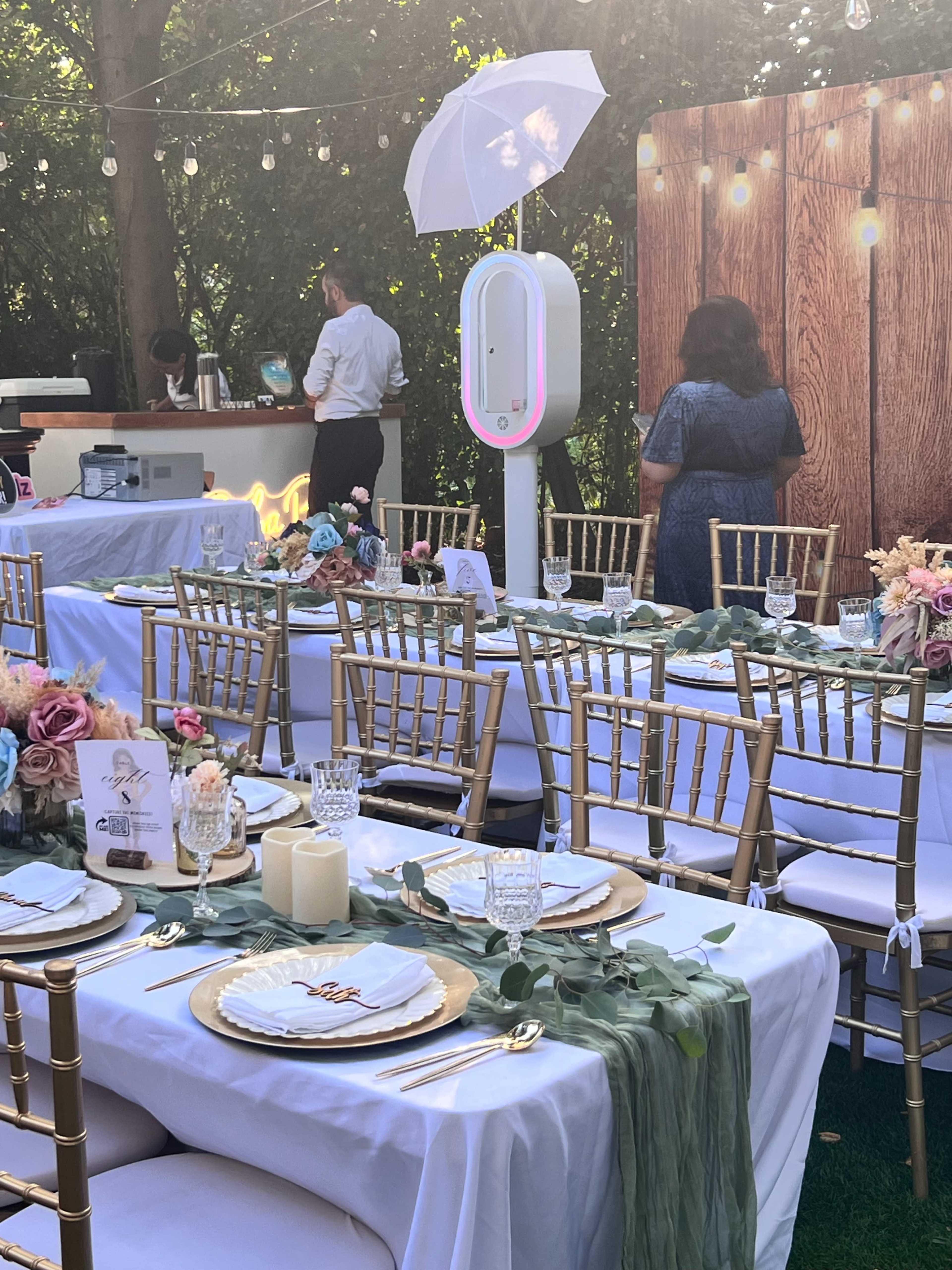 The scene depicts an elegantly arranged outdoor dining setup for an event, featuring tables adorned with white linens, golden chairs, and floral centerpieces, along with a backdrop of wooden panels and string lights.