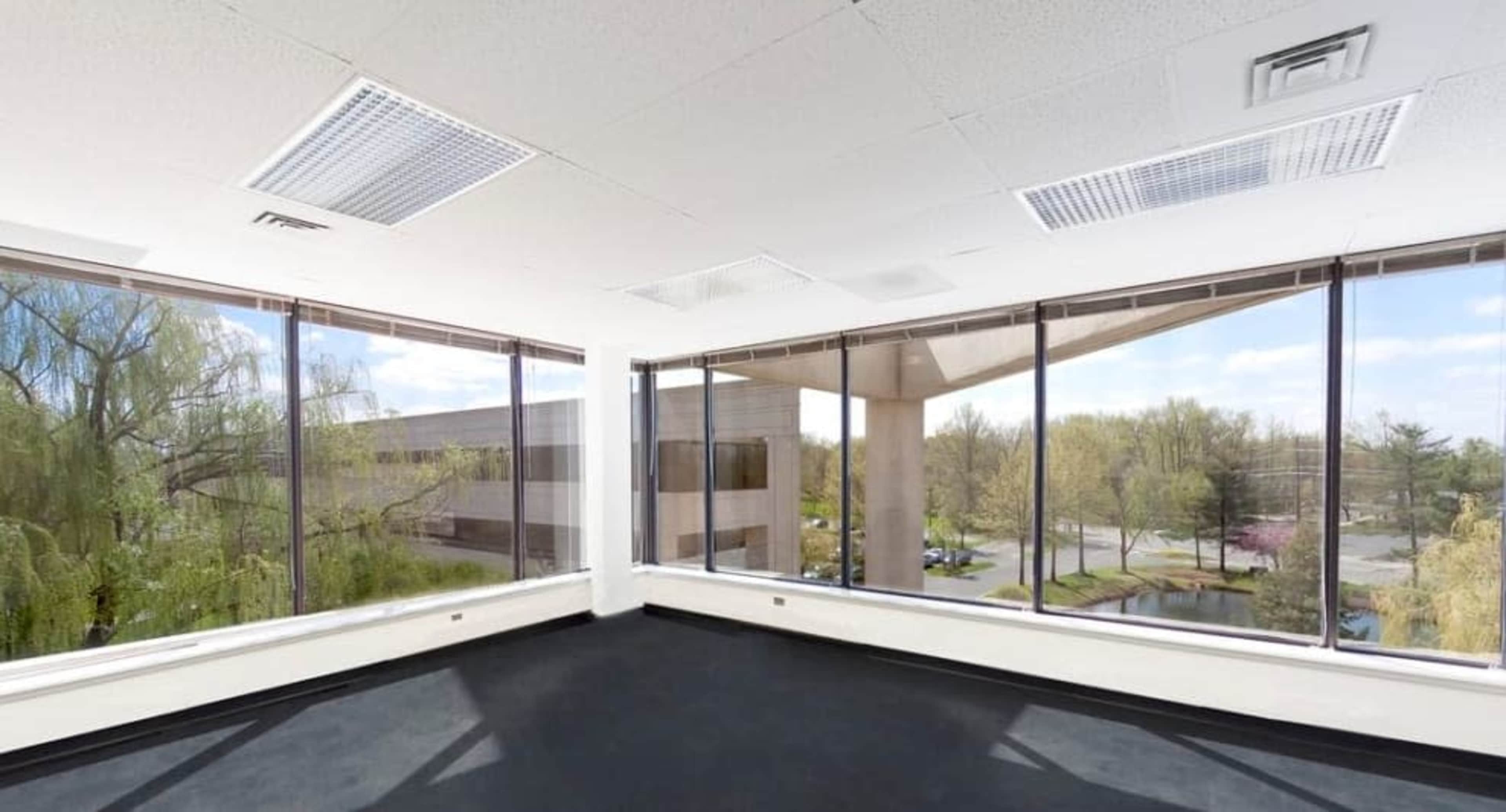 The image shows a vacant office space with large windows offering a view of the surrounding landscaped area outside.