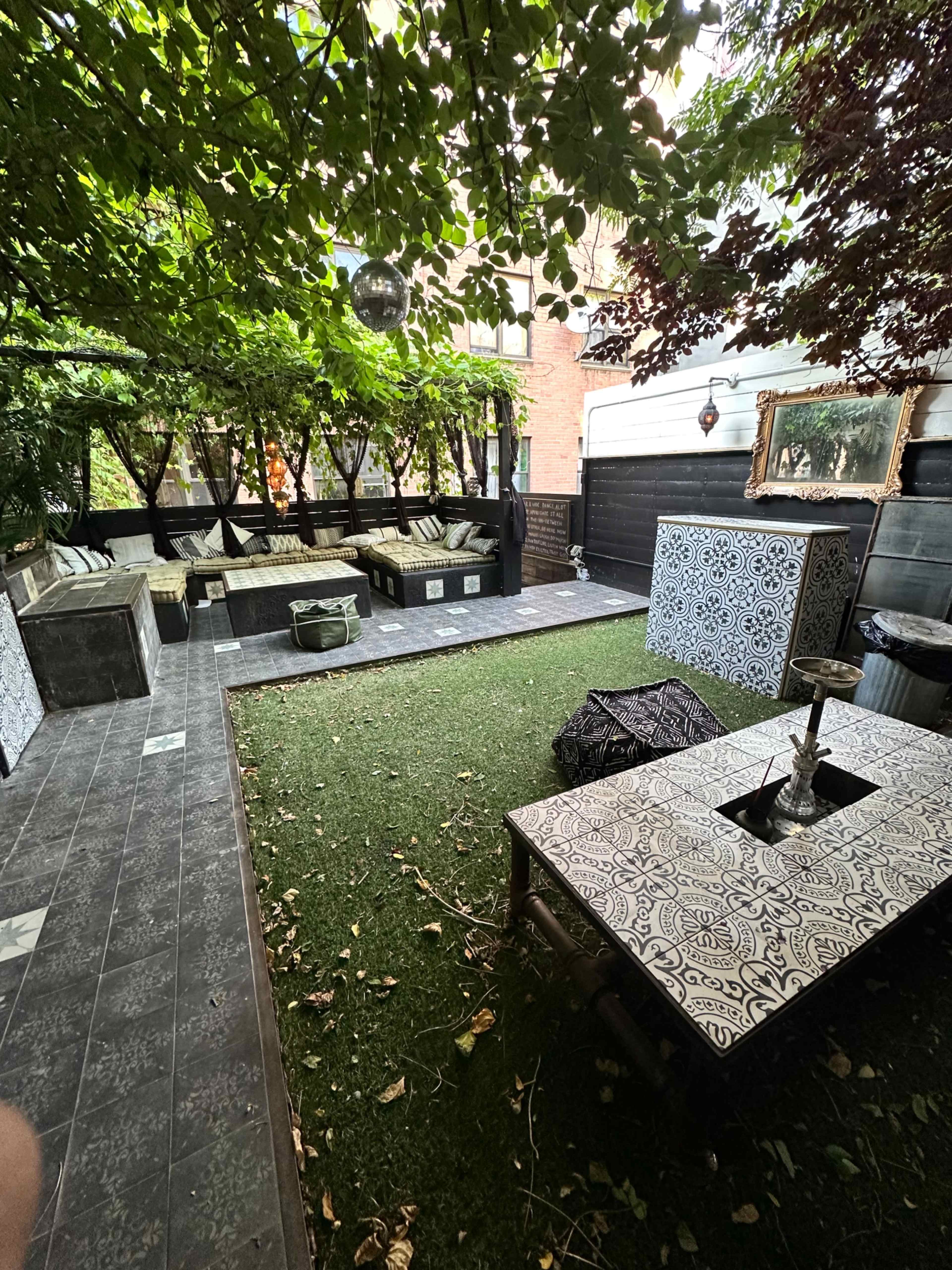 The image shows a landscaped outdoor space with seating areas, decorative tables, and greenery overhead.