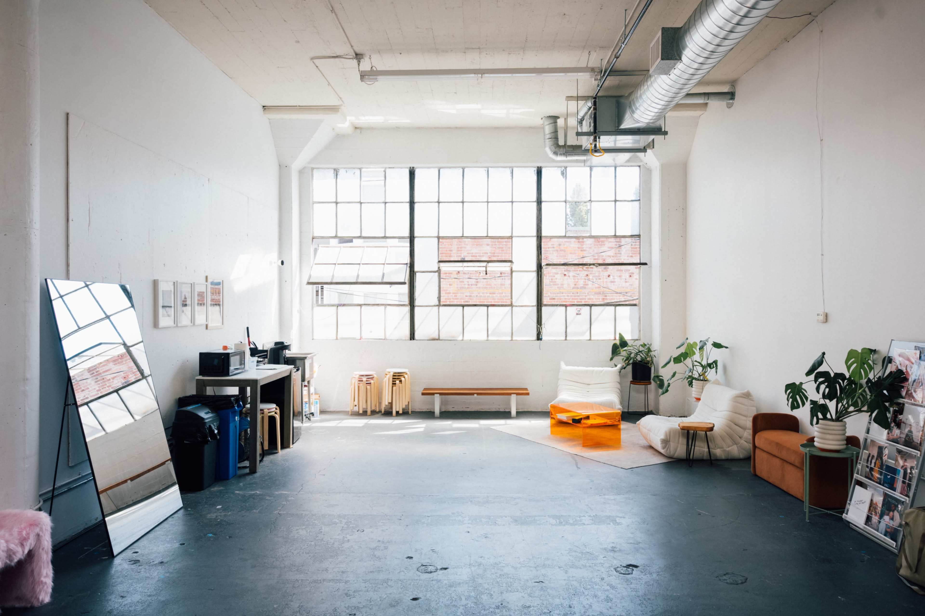 The image shows a spacious, brightly lit room with large windows, featuring minimalist furniture including a couch, a sitting area with cushions, and a work station against one wall.