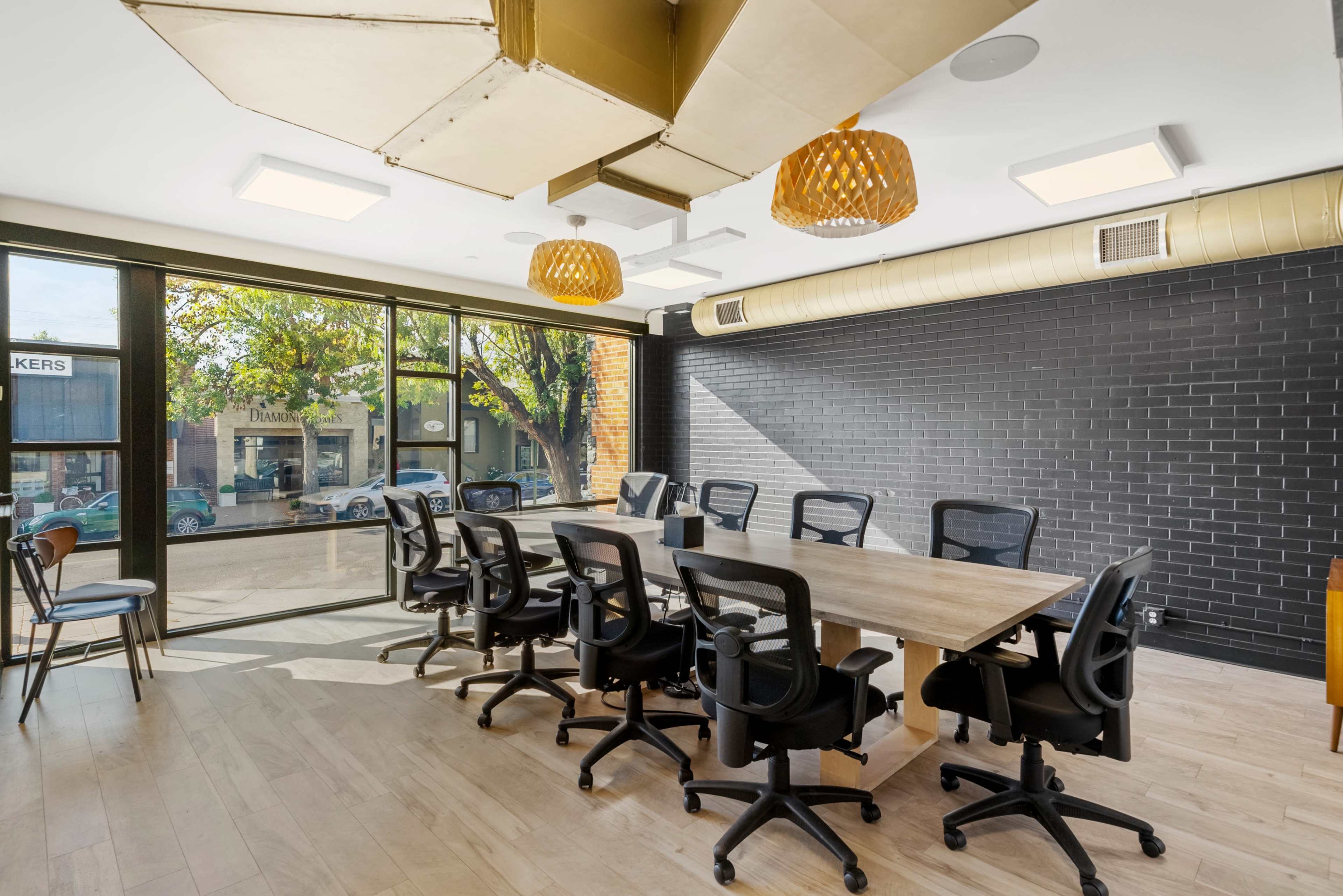 A modern conference room features a large wooden table surrounded by ergonomic chairs, with large windows providing a view of the street outside.