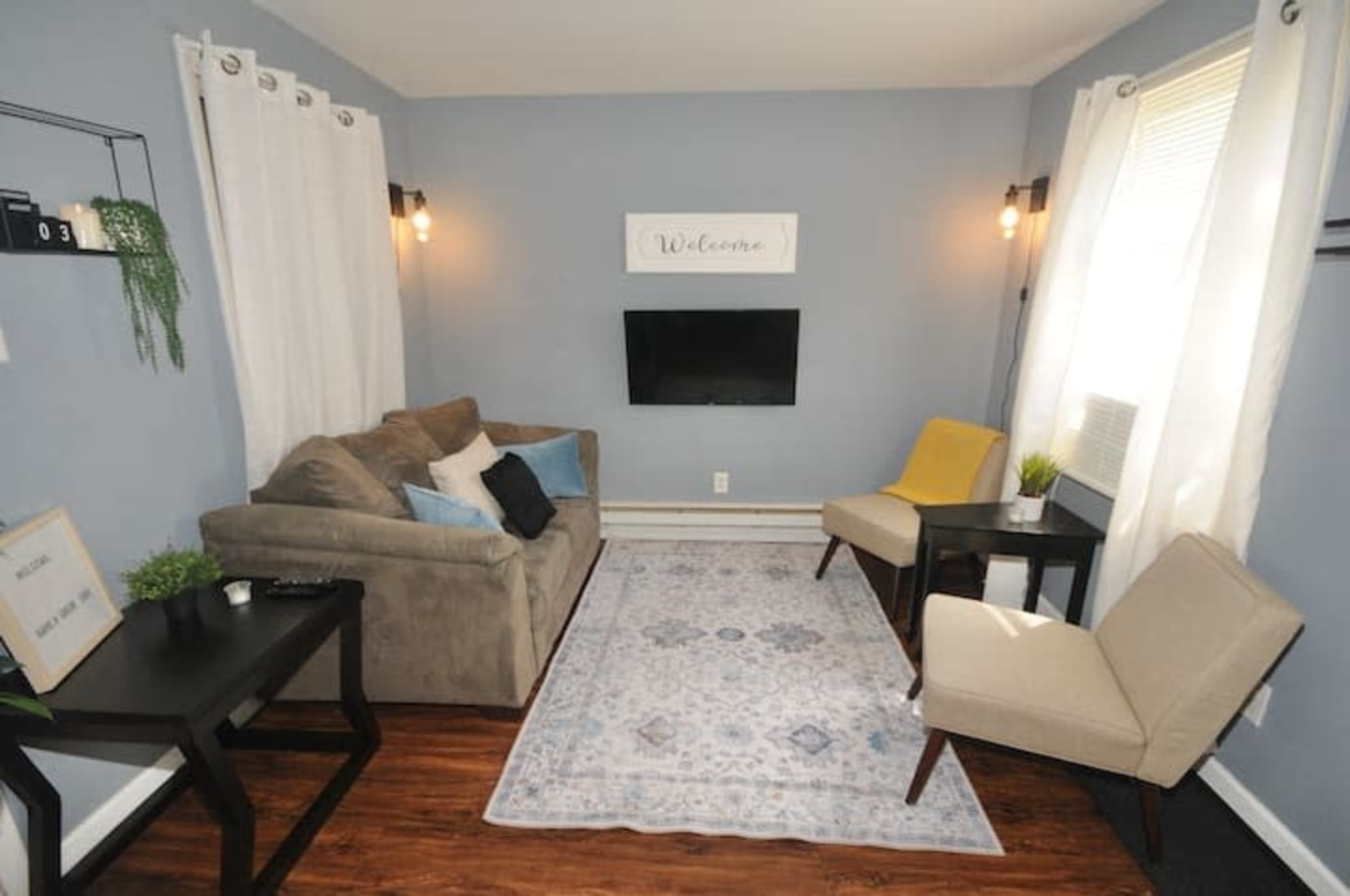A small living room with a gray sofa, two chairs, a television mounted on the wall, and a decorative rug on a wooden floor.