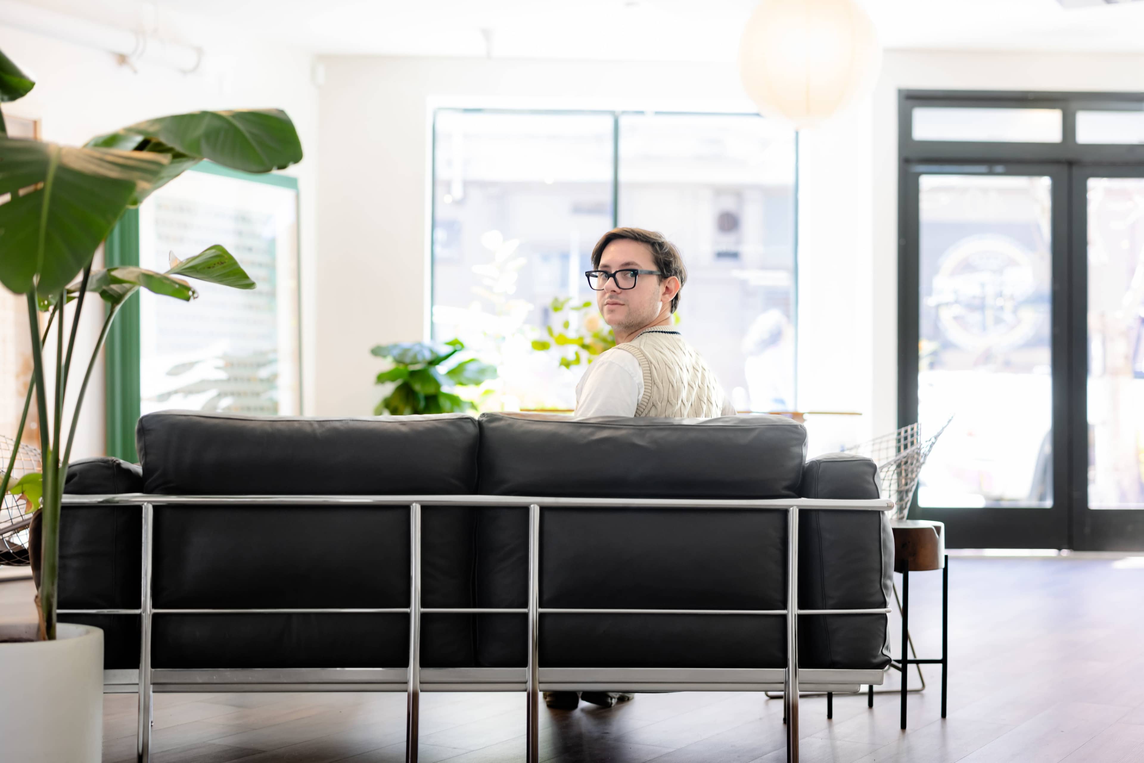 A person wearing glasses and a sweater vest sits on a modern black sofa, looking back toward the camera in a well-lit indoor space with plants and large windows.