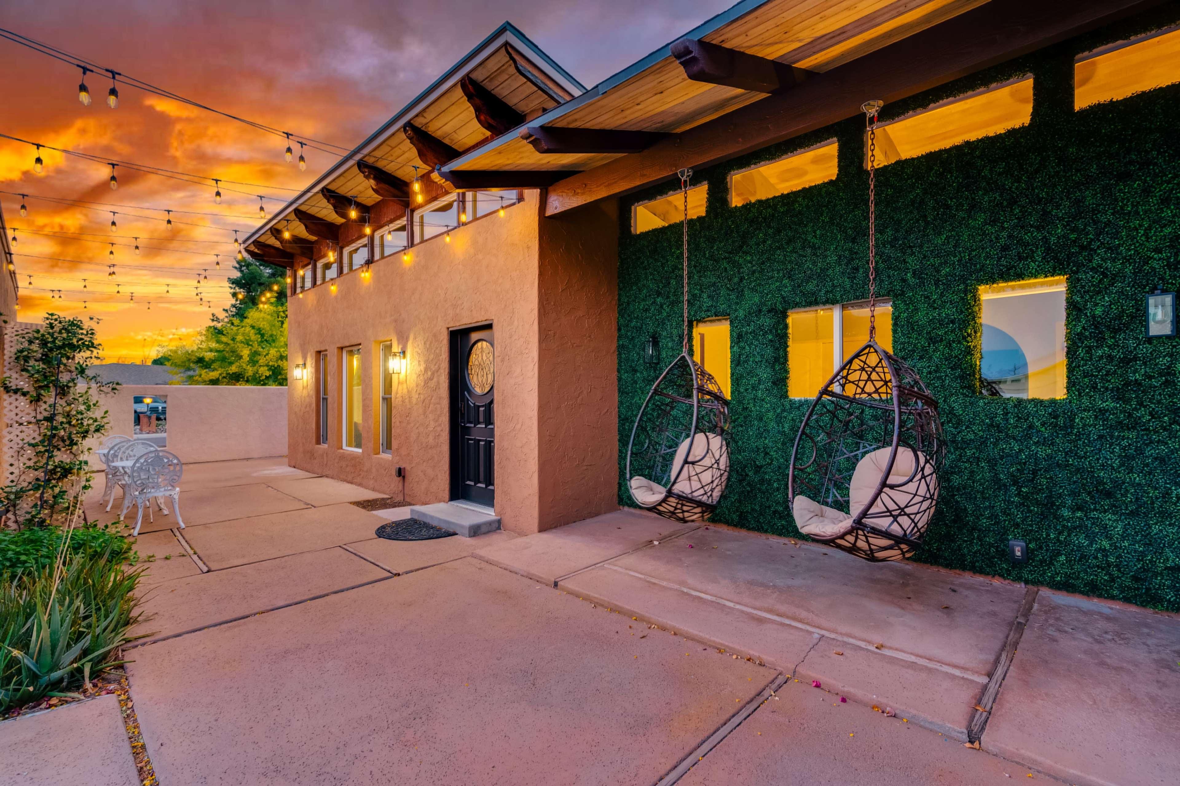A modern home features two hanging chairs beside a textured green wall, with a paved walkway leading to the entrance under a colorful sunset sky.