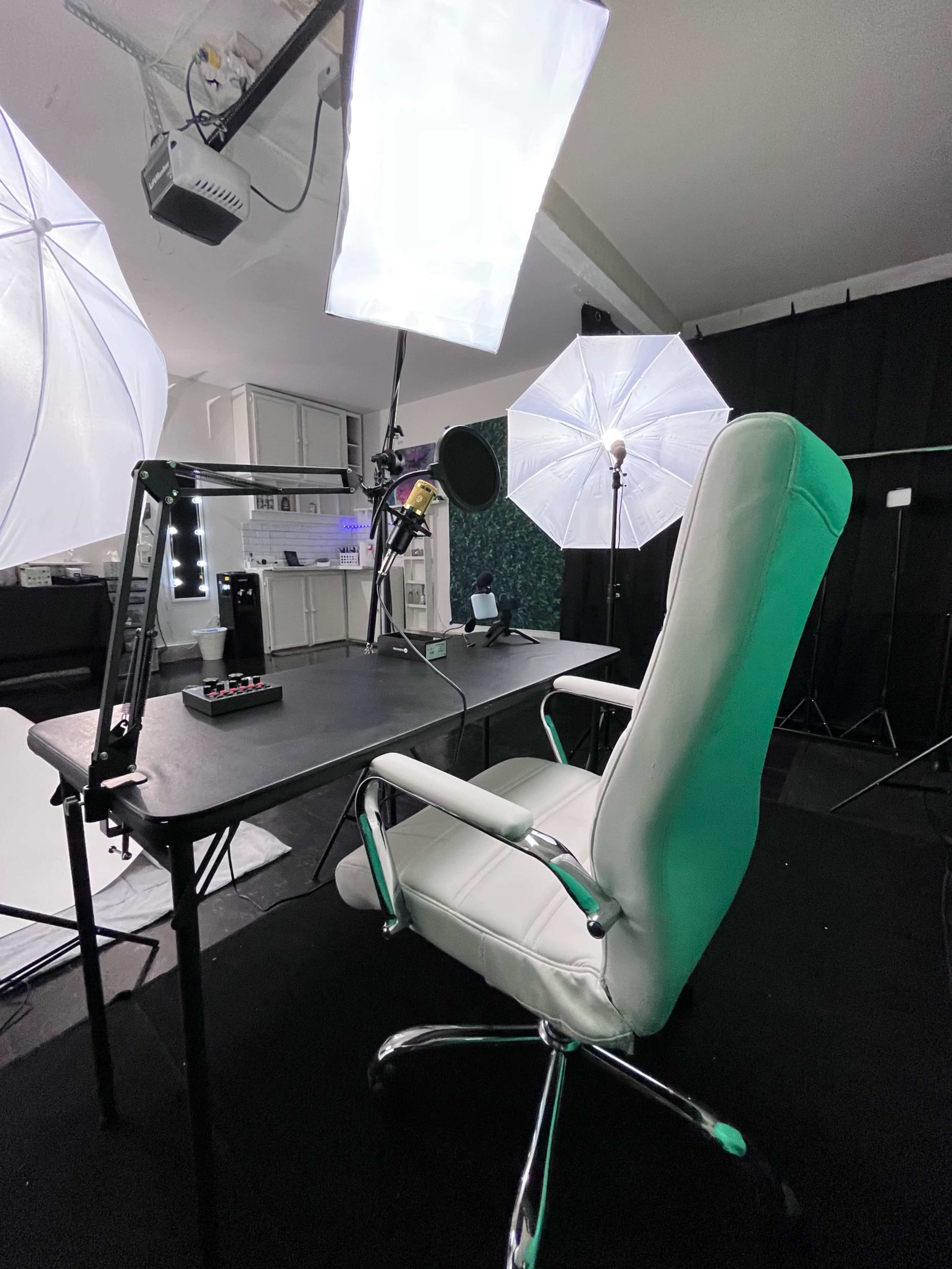 A white ergonomic office chair faces a black desk equipped with microphones and lighting equipment in a studio setup.