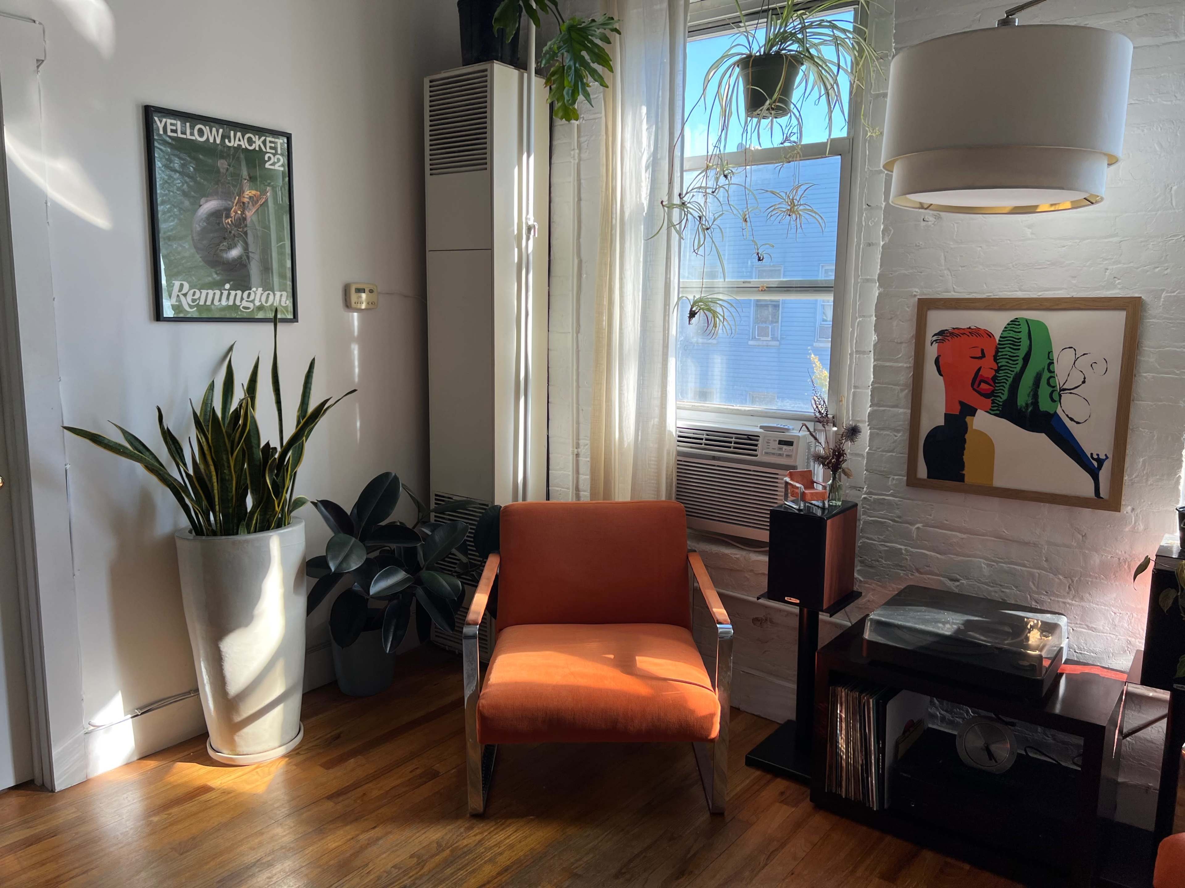 A vibrant living space features an orange armchair next to a window, surrounded by plants and a framed artwork on the wall.