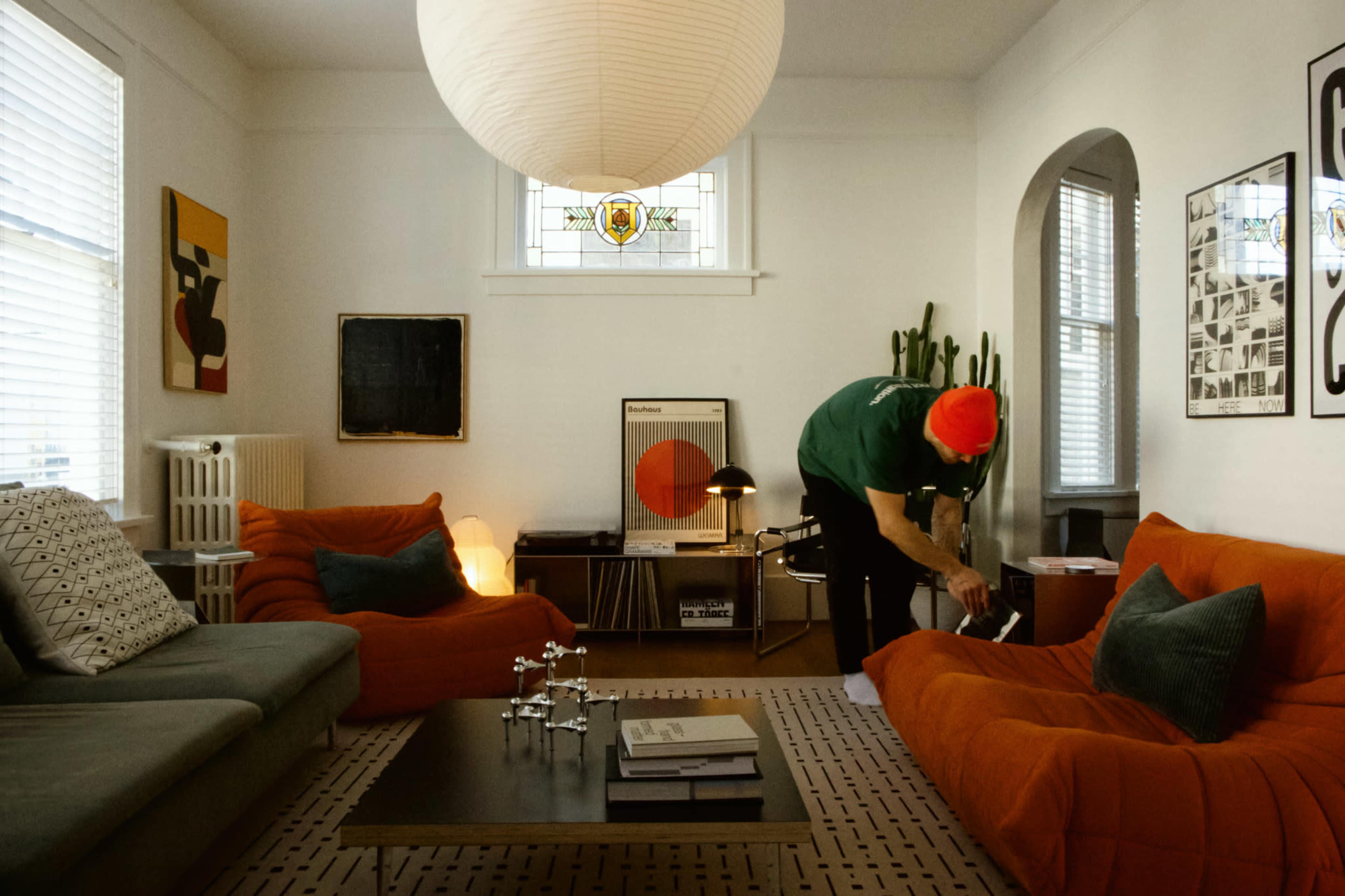 A person in a green shirt and red beanie is arranging items on an orange couch in a modern living room adorned with artwork and plants.