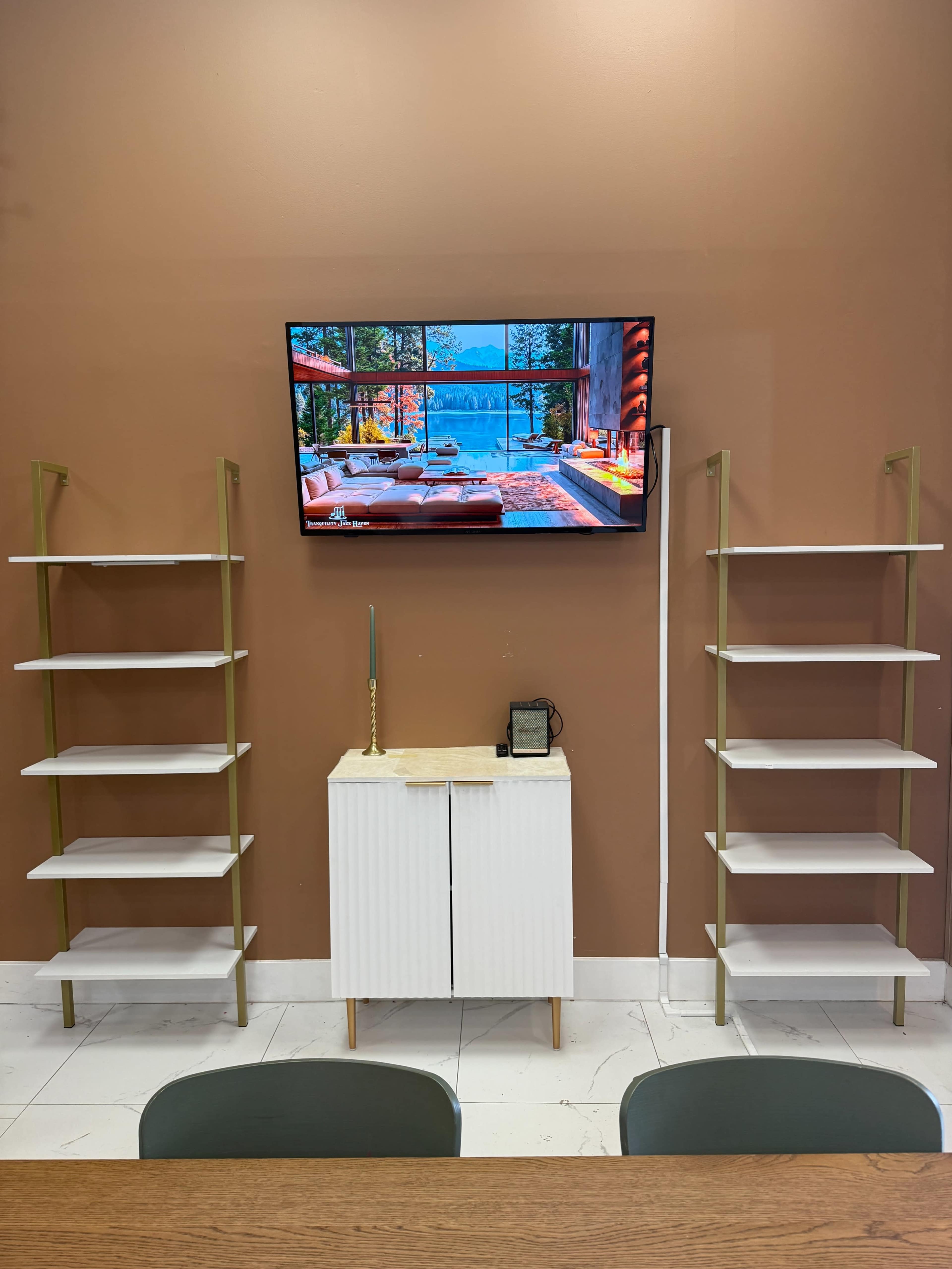 The image shows a modern interior featuring a television mounted above a white cabinet, flanked by two tall, gold-framed shelving units, against a brown wall.