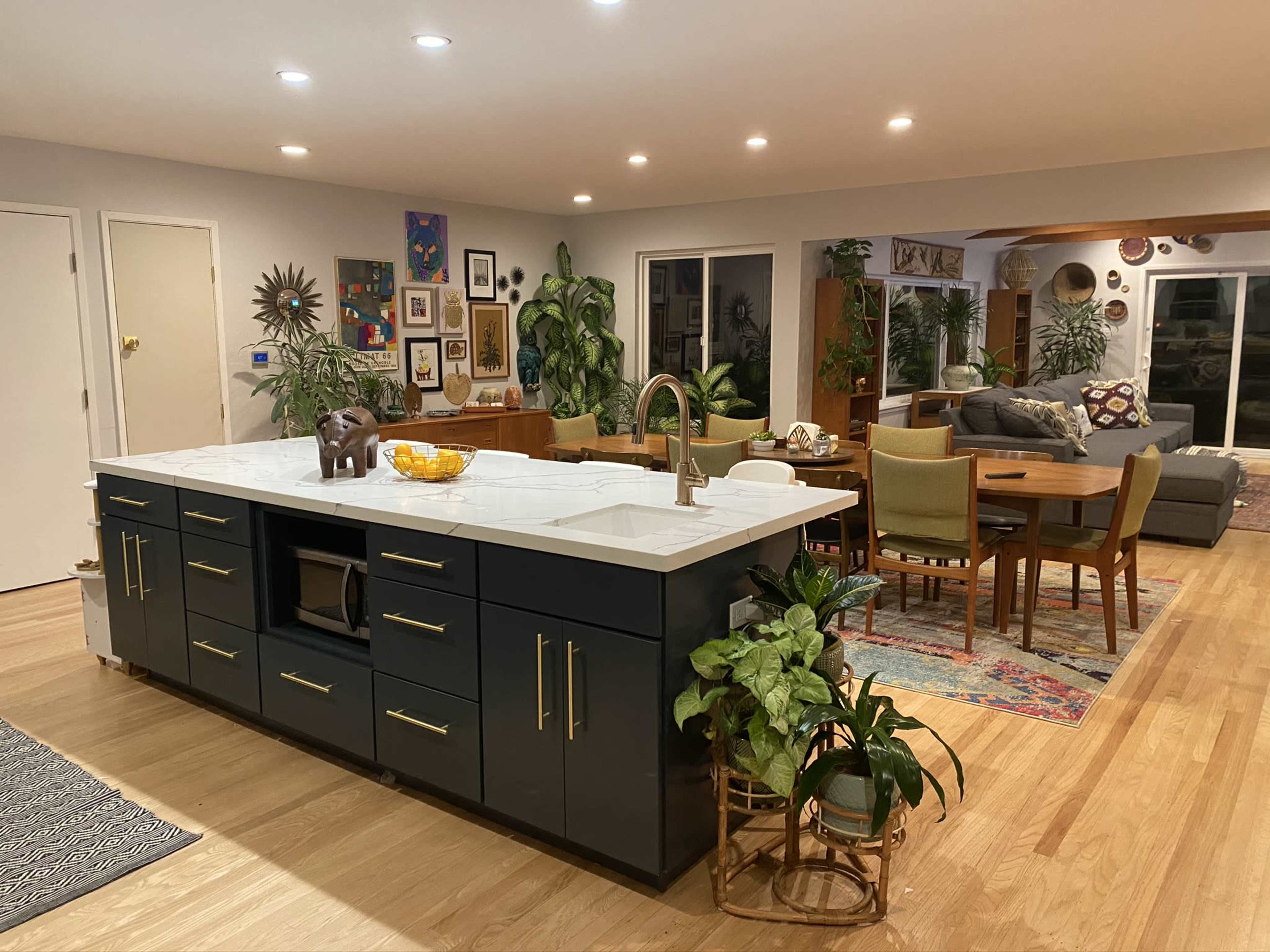 A modern kitchen with an island at the center, surrounded by dining and living areas featuring plants, artwork, and various furnishings.