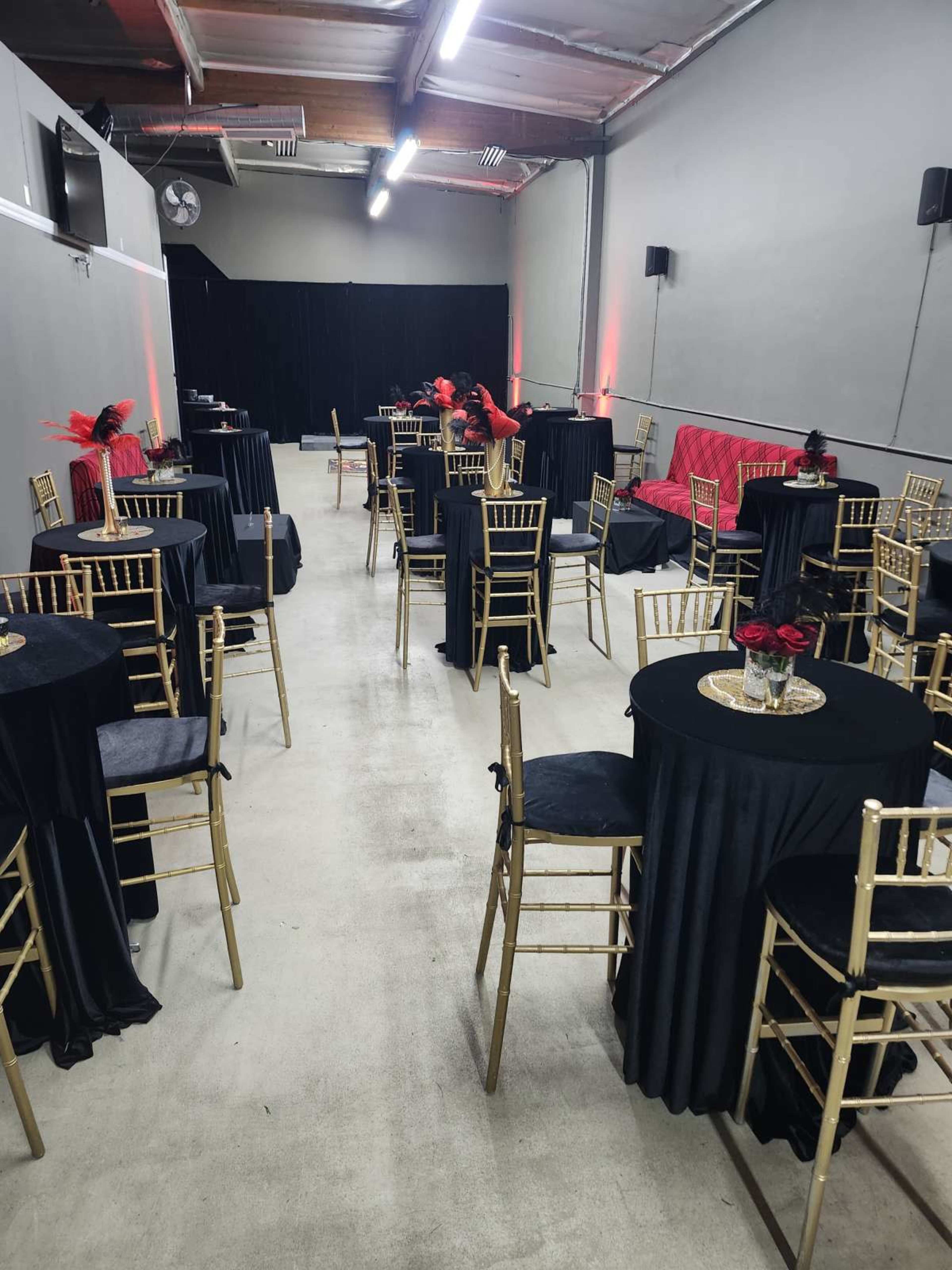 The image shows a partially set up event space with several round tables covered in black cloth, surrounded by gold chairs, and decorated with red floral arrangements.