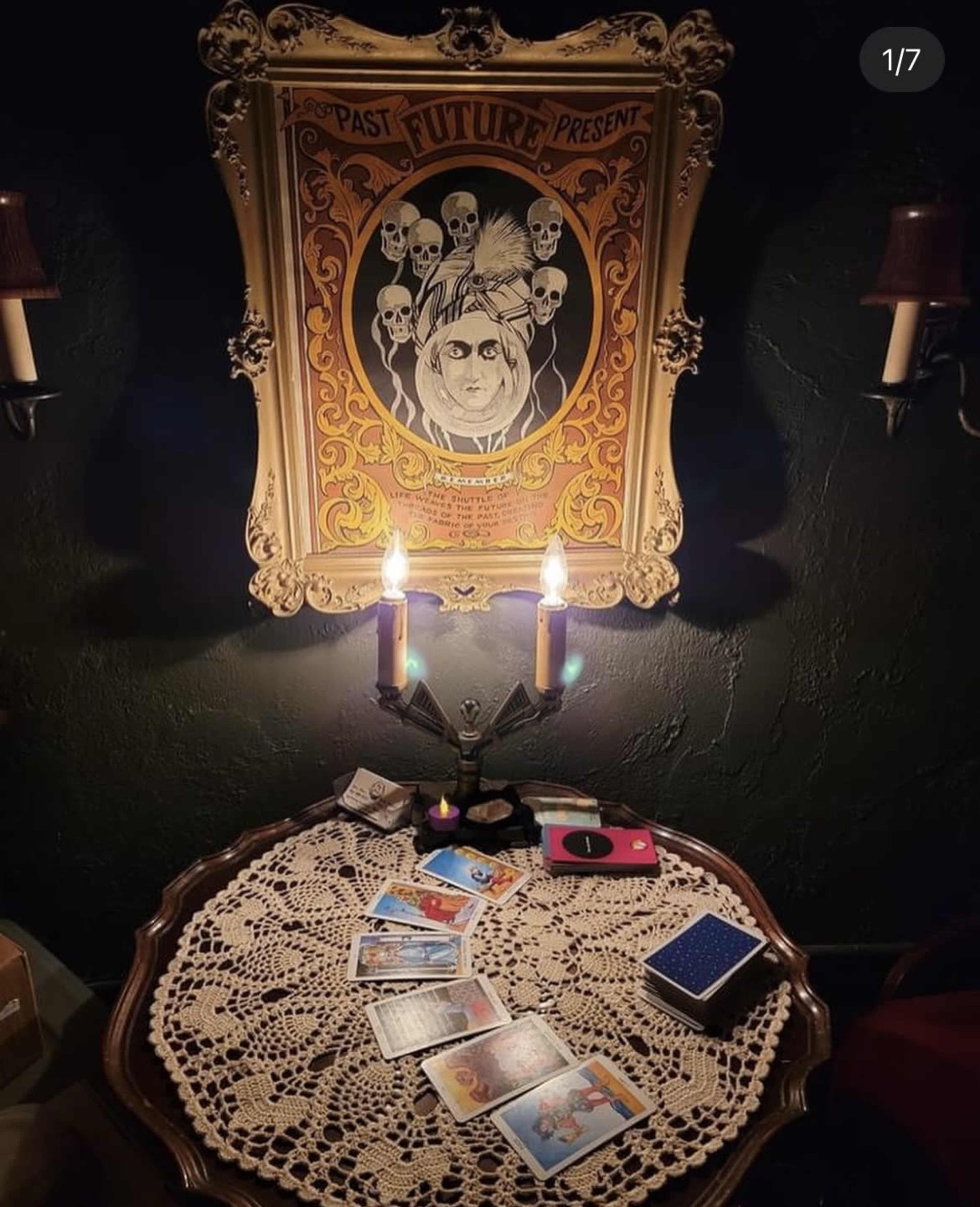 A dimly lit room featuring a round table with a lace tablecloth, tarot cards scattered on it, and a framed poster of skulls hanging on the wall above a pair of lit candles.
