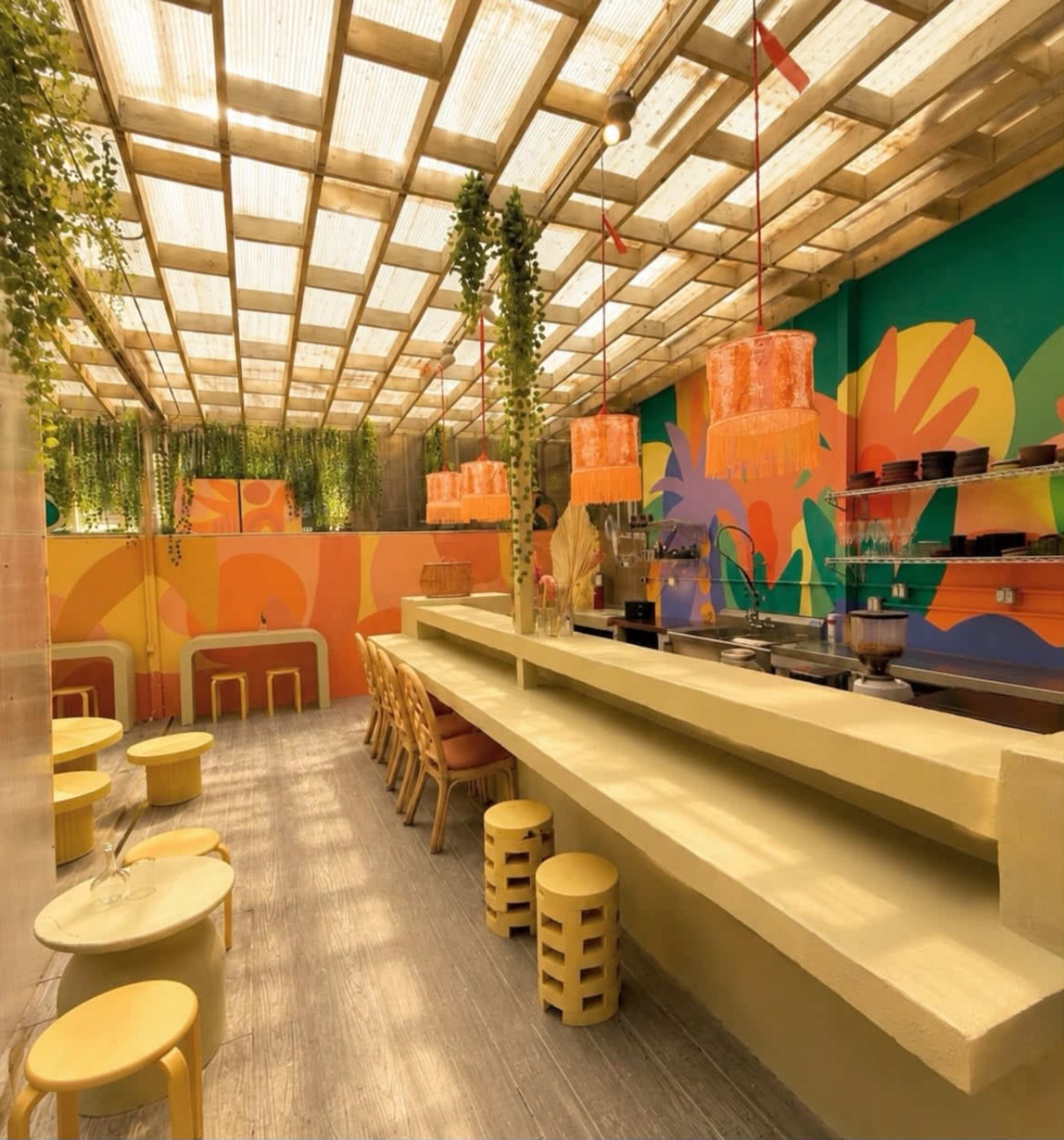 The image shows a brightly colored indoor restaurant with a bar area, featuring wooden stools and vibrant wall art, illuminated by natural light filtering through a transparent roof.