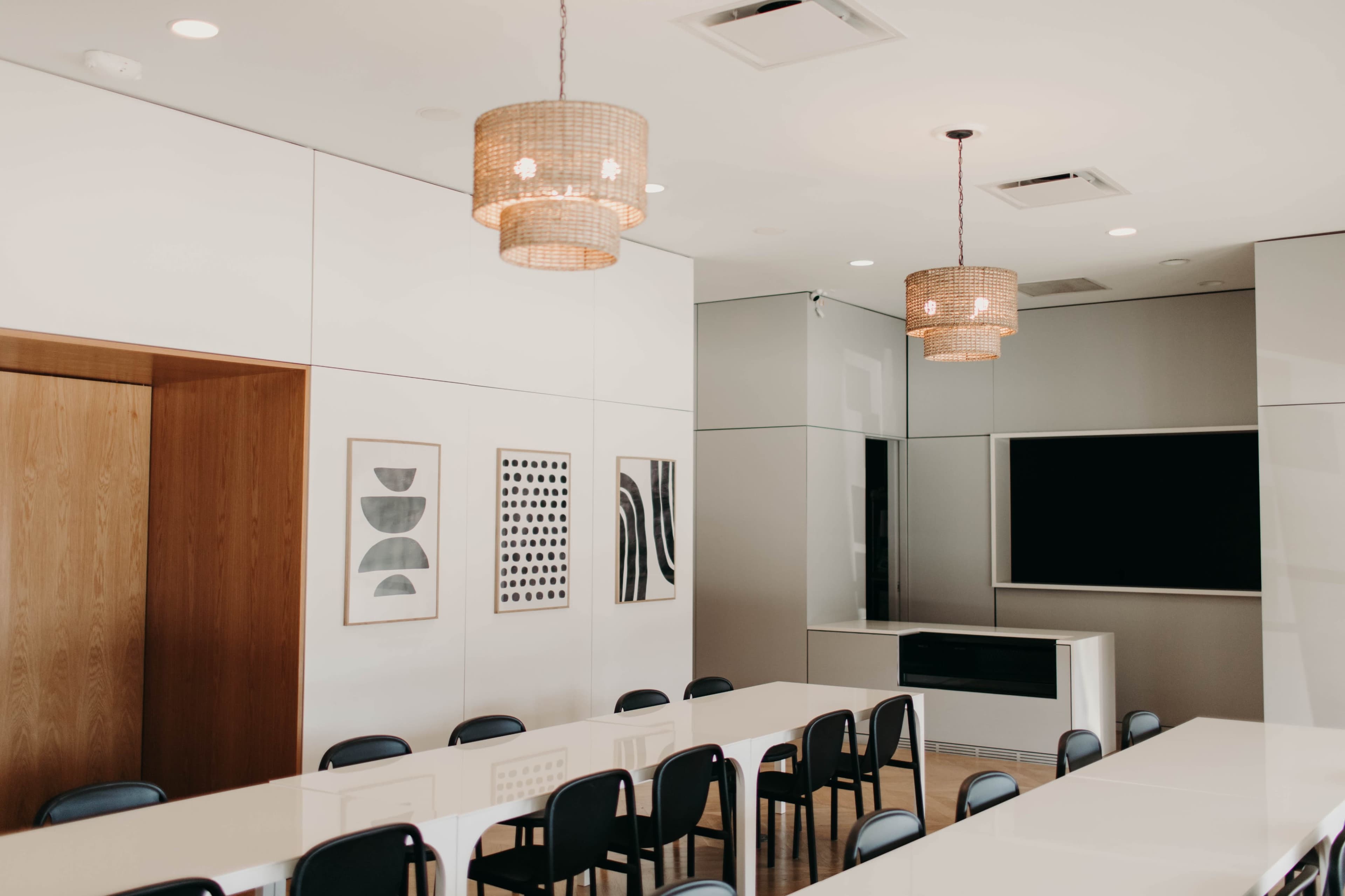 A modern conference room featuring a long white table, black chairs, and framed artwork on the walls, illuminated by woven pendant lights.