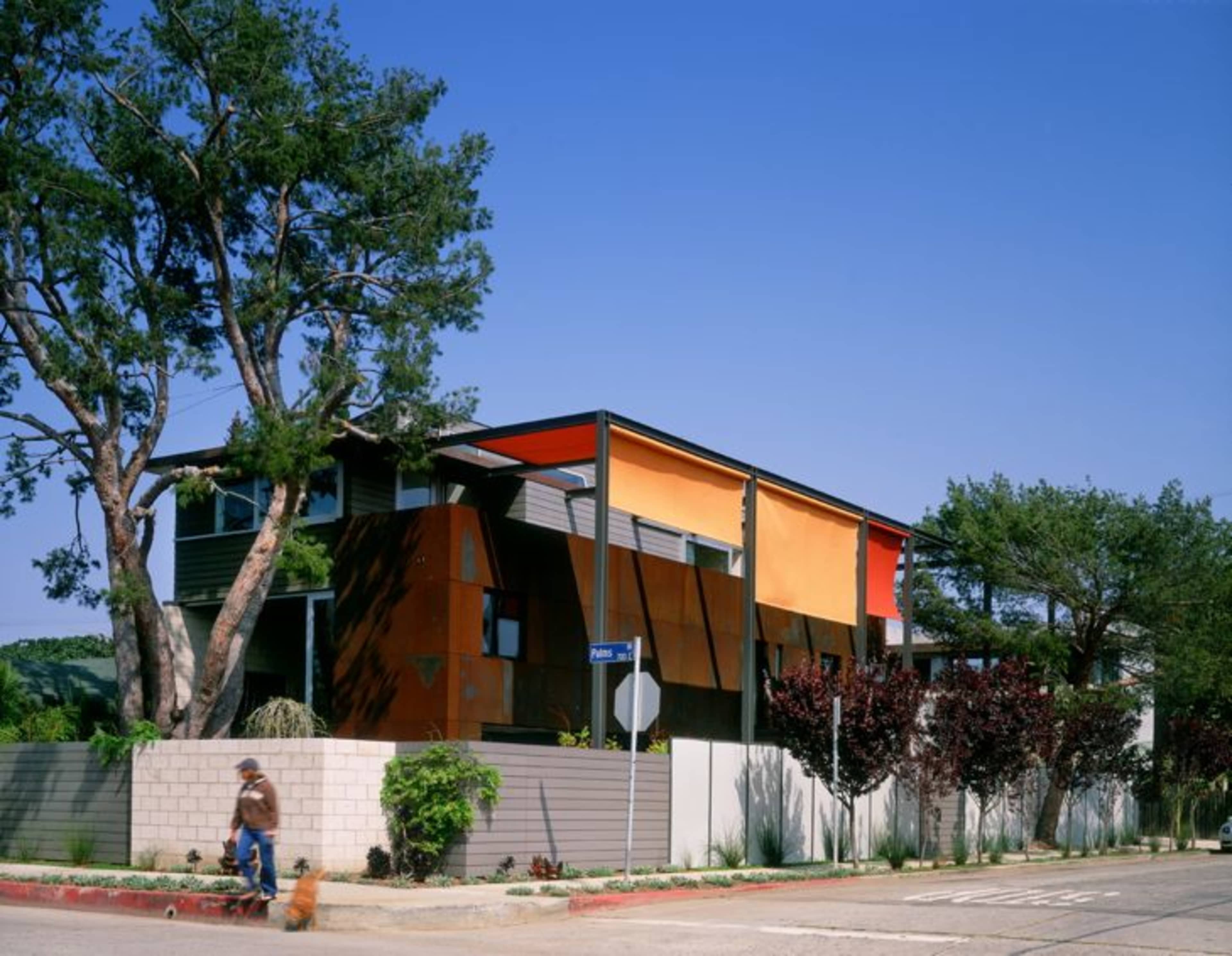 A modern house with orange and gray exterior panels is surrounded by trees and a low wall, with a person walking a dog along the sidewalk.