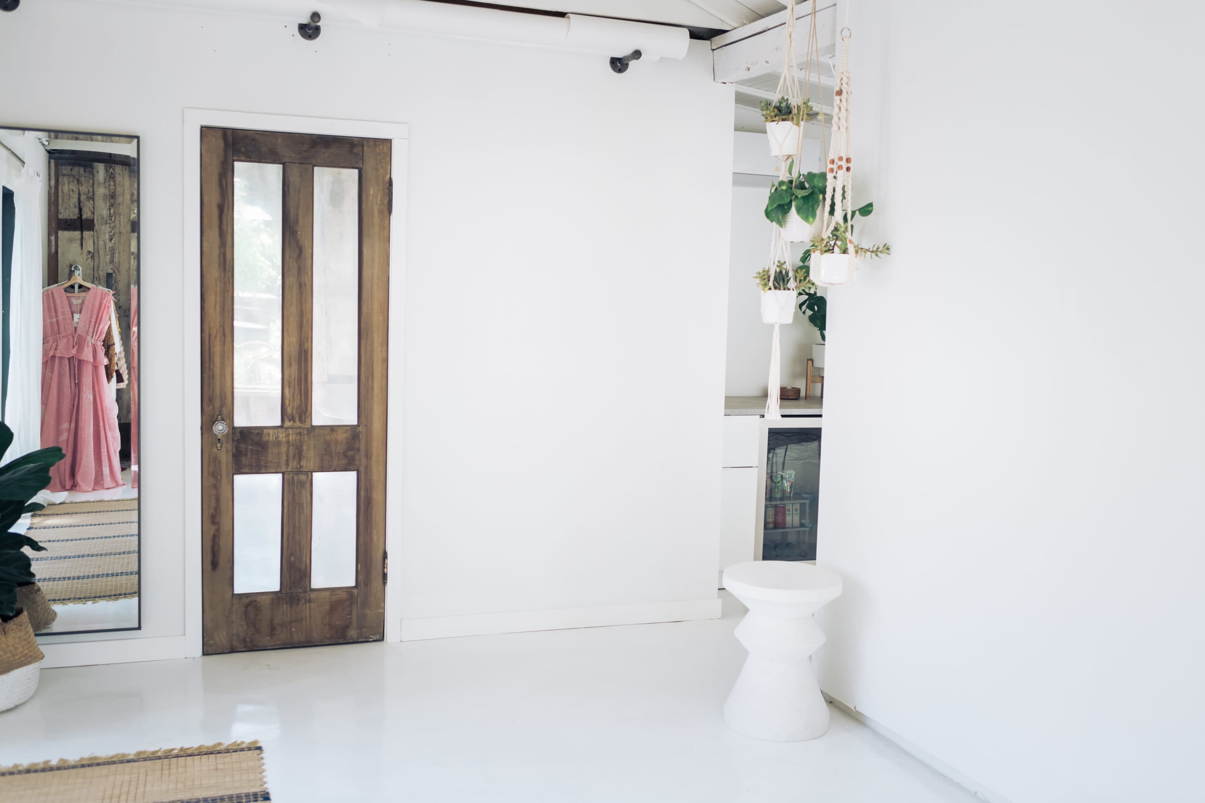The image shows a bright, minimalist interior featuring a wooden door, a small white stool, and hanging planters, with a glimpse of a kitchen area and a dress displayed on a rack.