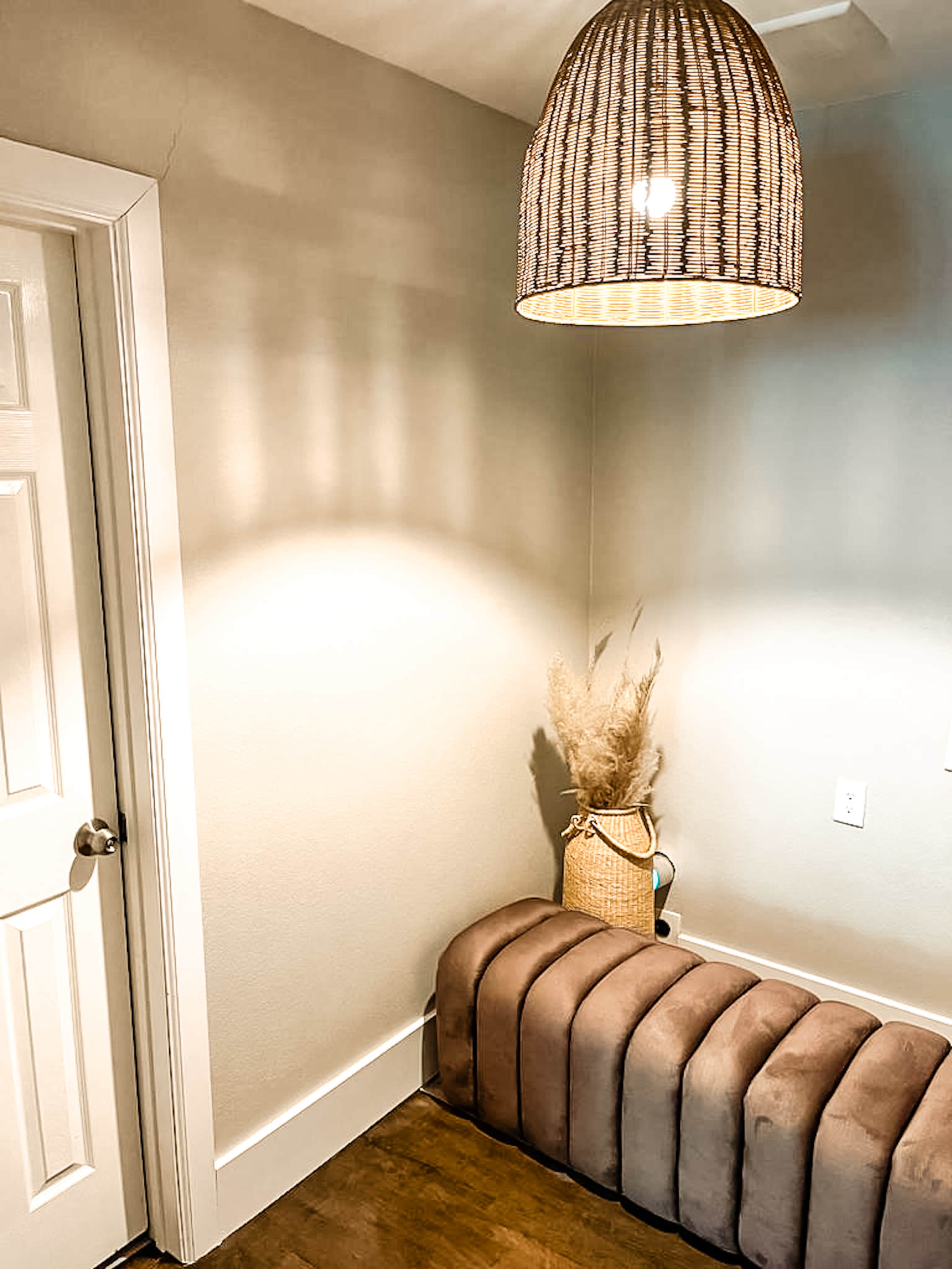 A corner of a room features a woven pendant light above a brown padded bench and a decorative basket with dried plants.