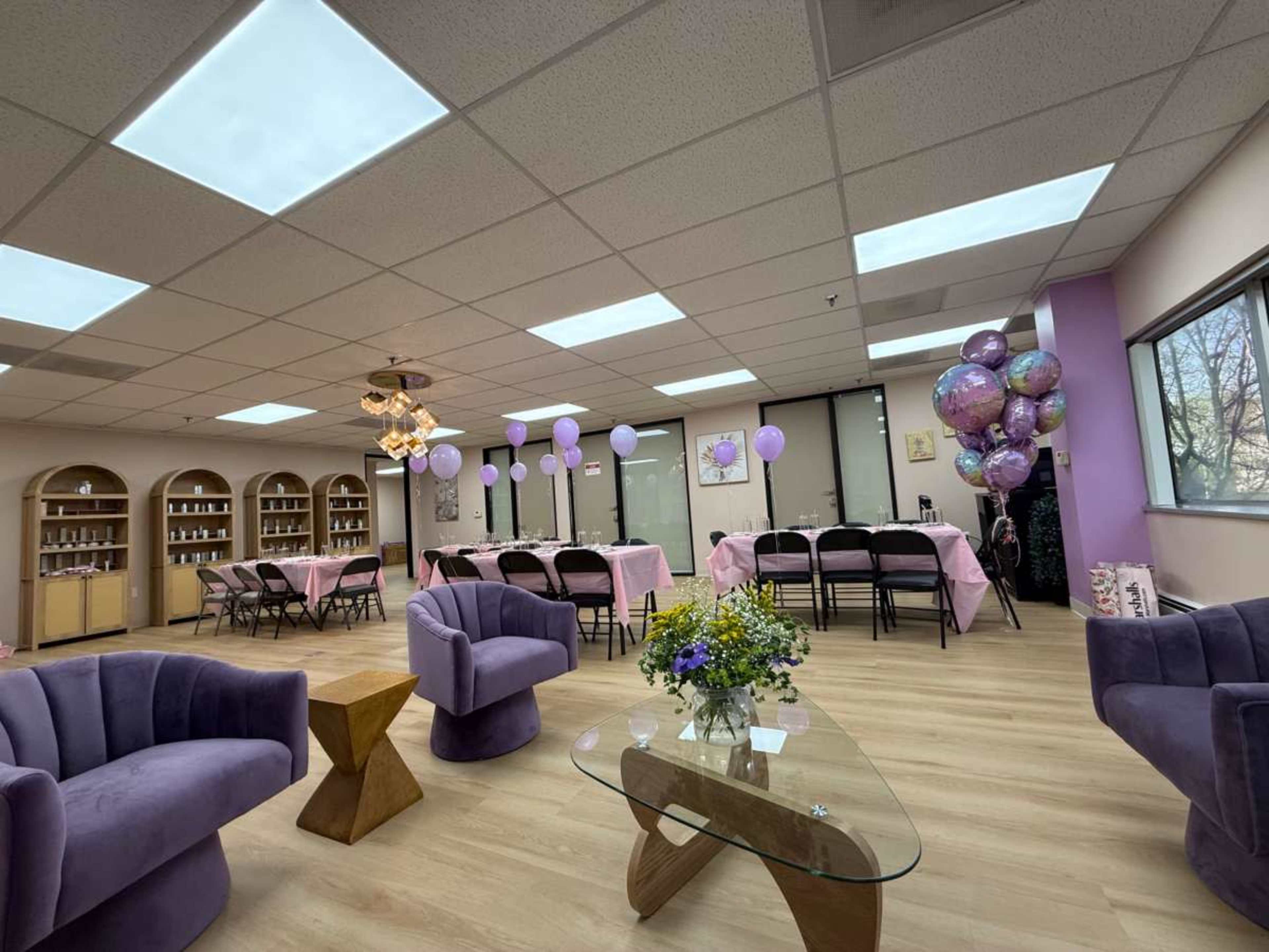The image shows a decorated interior space with purple balloons and tables set with pink linens, accompanied by modern seating arrangements and shelves in the background.