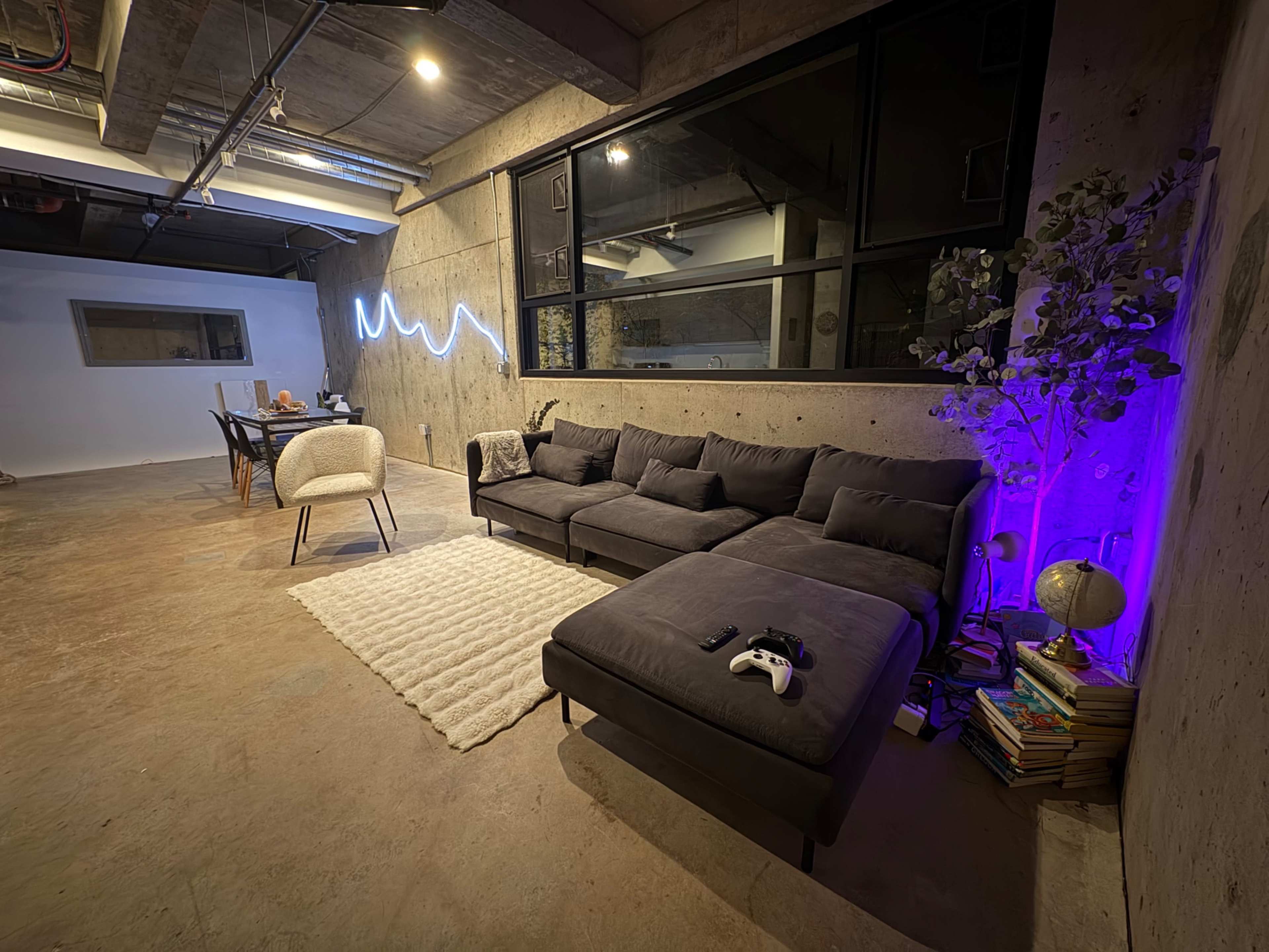 The image shows a modern, minimalistic living space featuring a gray sectional couch, a light rug, a small dining area, and ambient lighting along the walls.