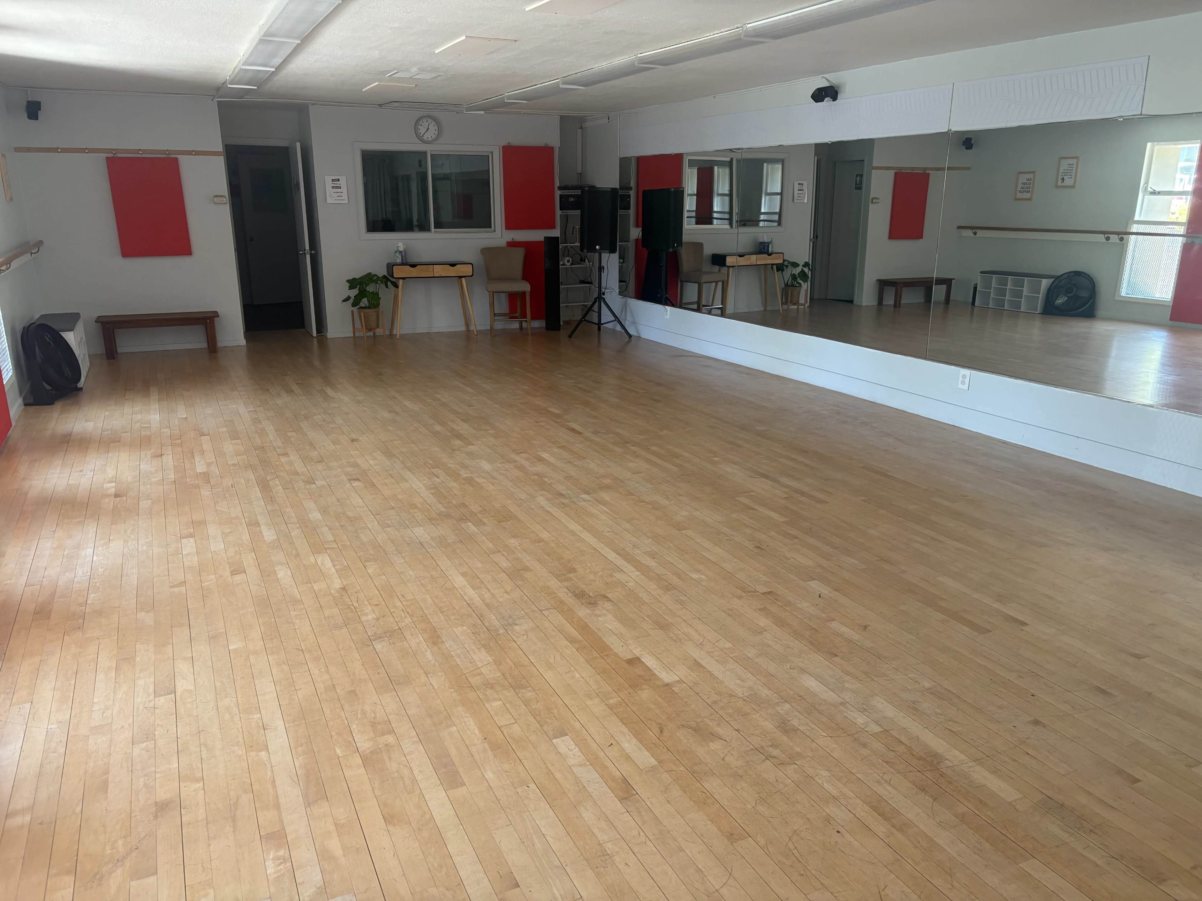 The image shows an empty dance studio with wooden flooring, mirrors on one wall, and a few pieces of furniture positioned around the room.