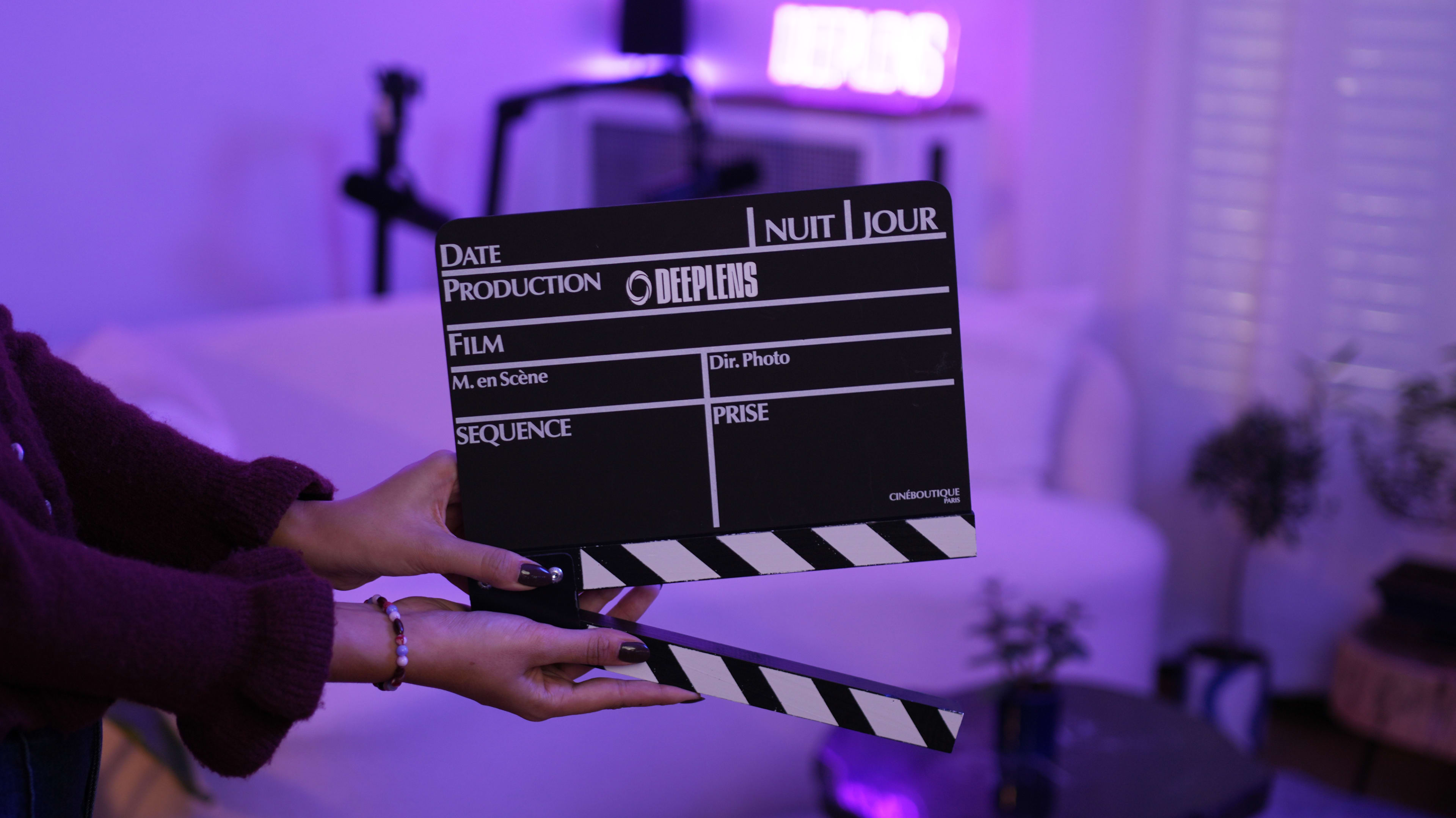 A person holds a film clapperboard in a purple-lit room with a white couch in the background.