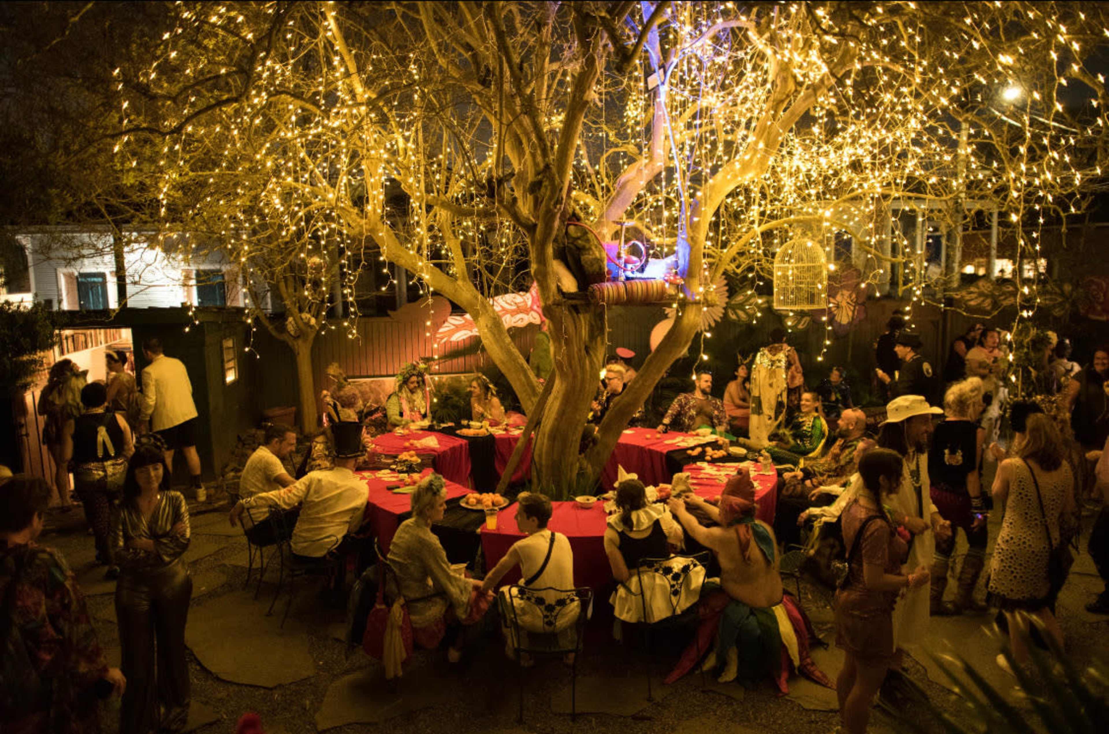 A large, festive gathering is taking place outdoors under a tree adorned with twinkling lights, featuring round tables filled with guests enjoying food and conversation.