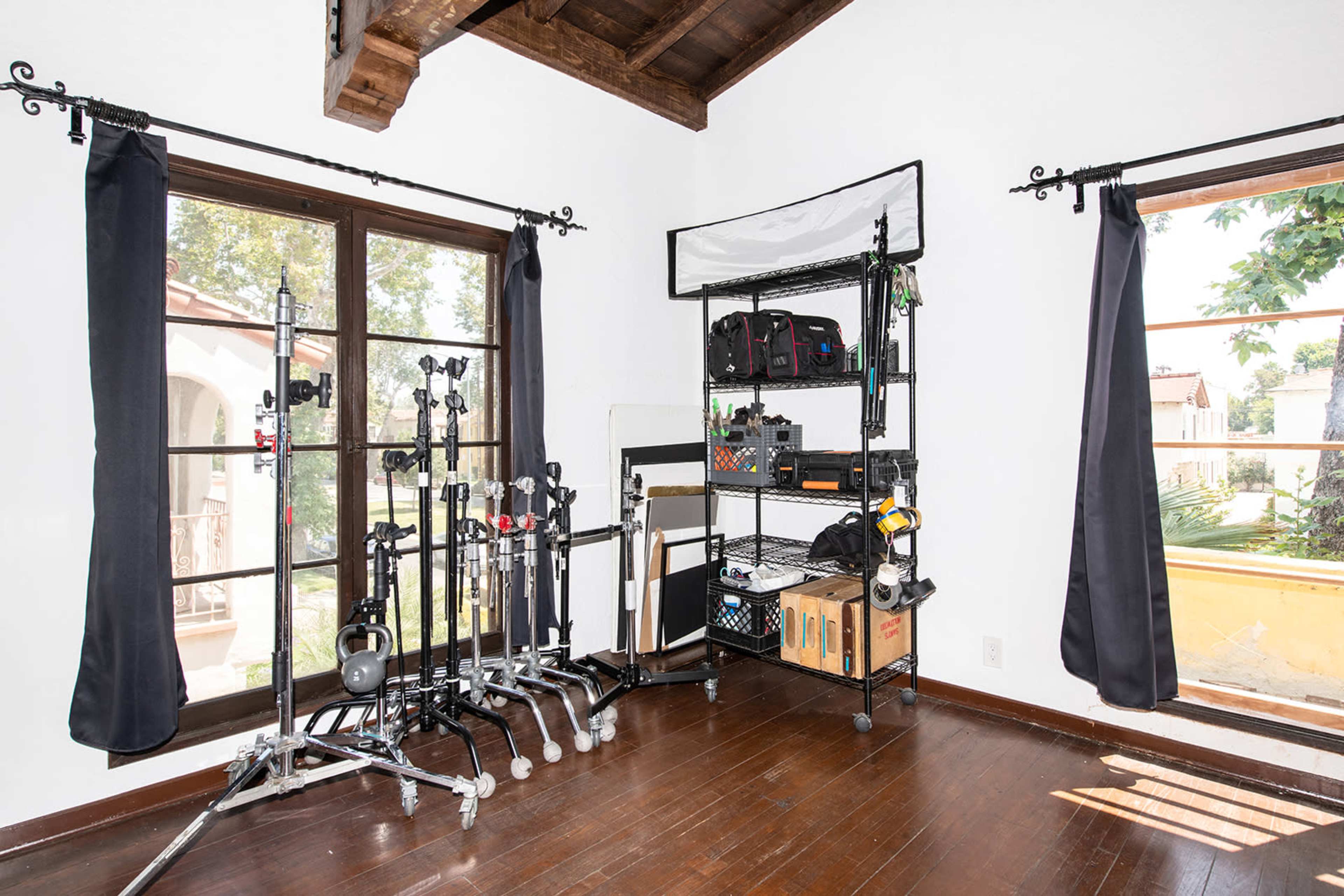 A room features two large windows with black curtains, a shelving unit filled with camera equipment, and several light stands arranged against the wall.