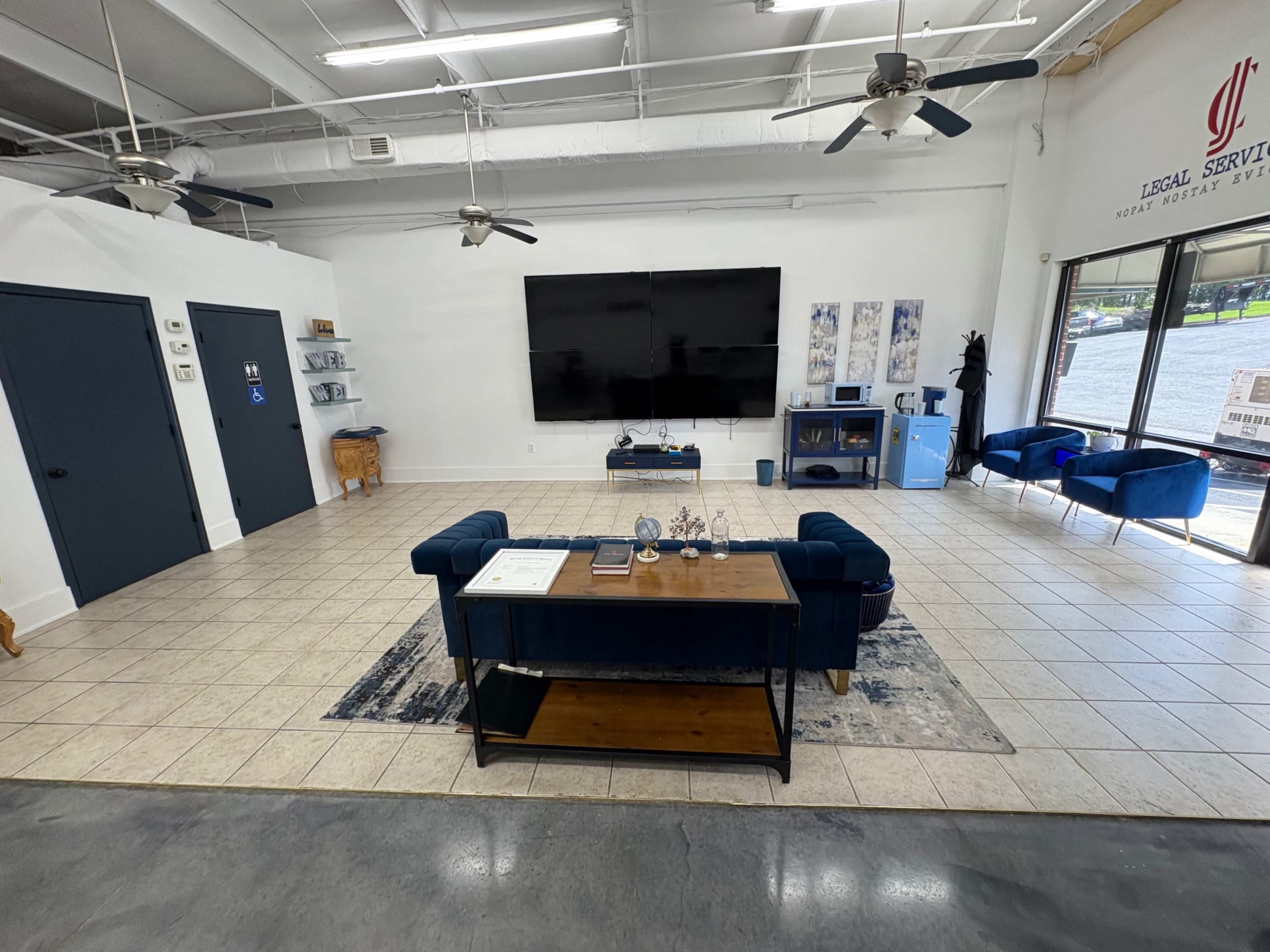 The image shows a modern office space with a large television mounted on the wall, a coffee table in the center, and seating arrangements consisting of blue furniture.