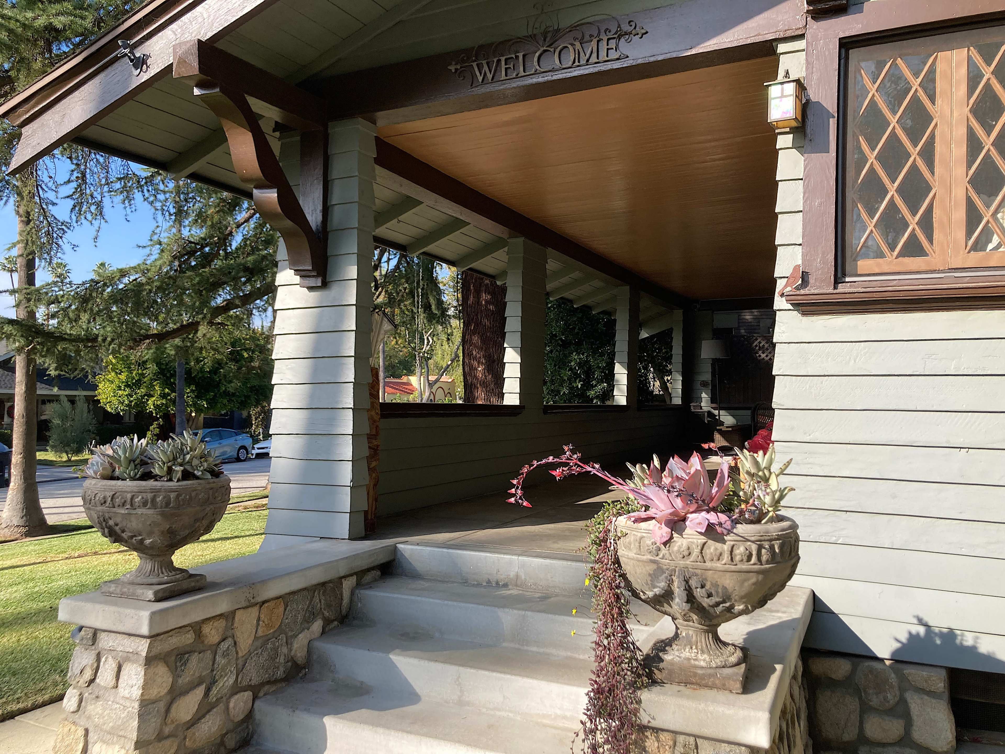 A front porch features stone steps and decorative planters with various plants, along with a "WELCOME" sign above.