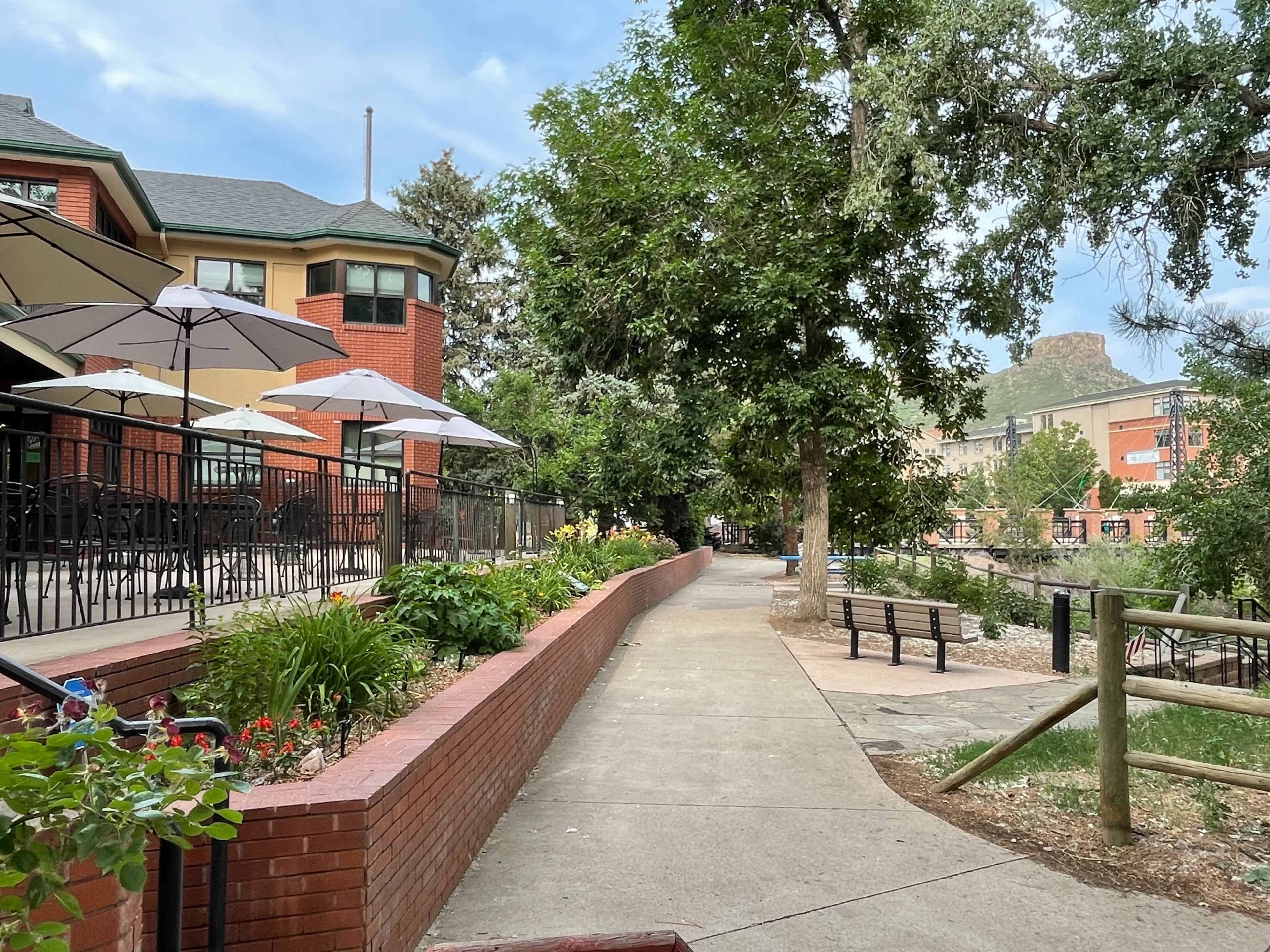 A paved walkway with landscaped flower beds runs alongside a building with outdoor seating under umbrellas.