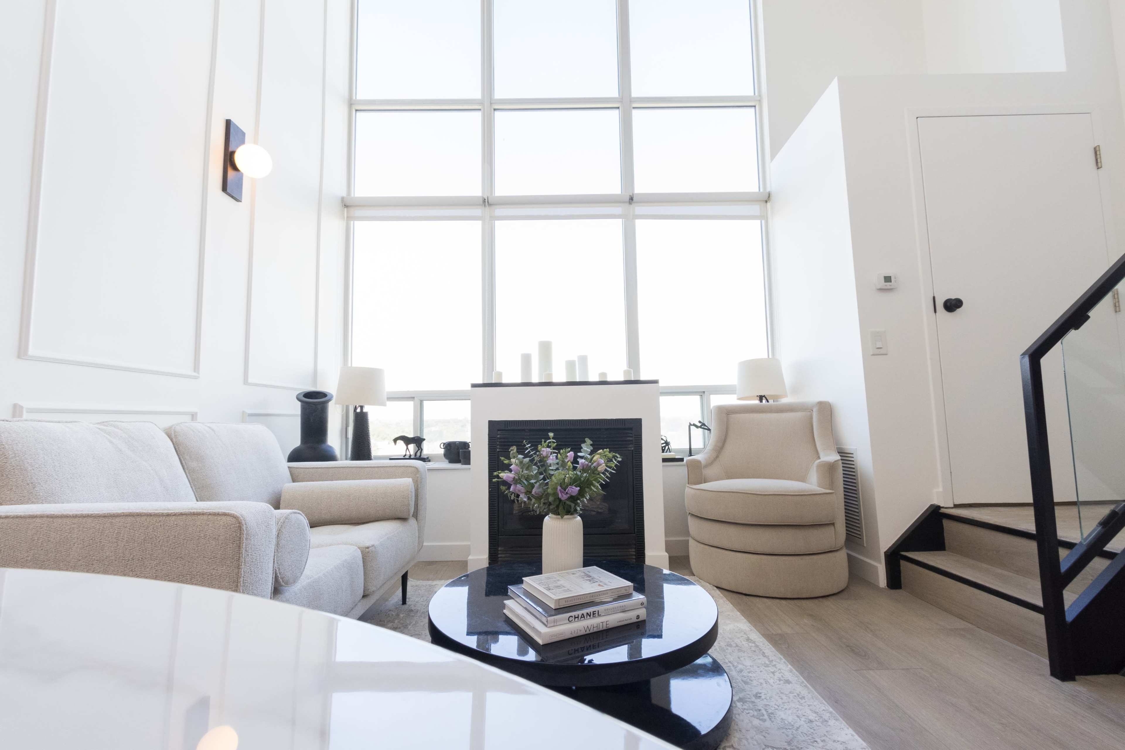 A modern living room features a light-colored sofa, a round coffee table, and a tall window with a view, complemented by a decorative fireplace and a cozy armchair.